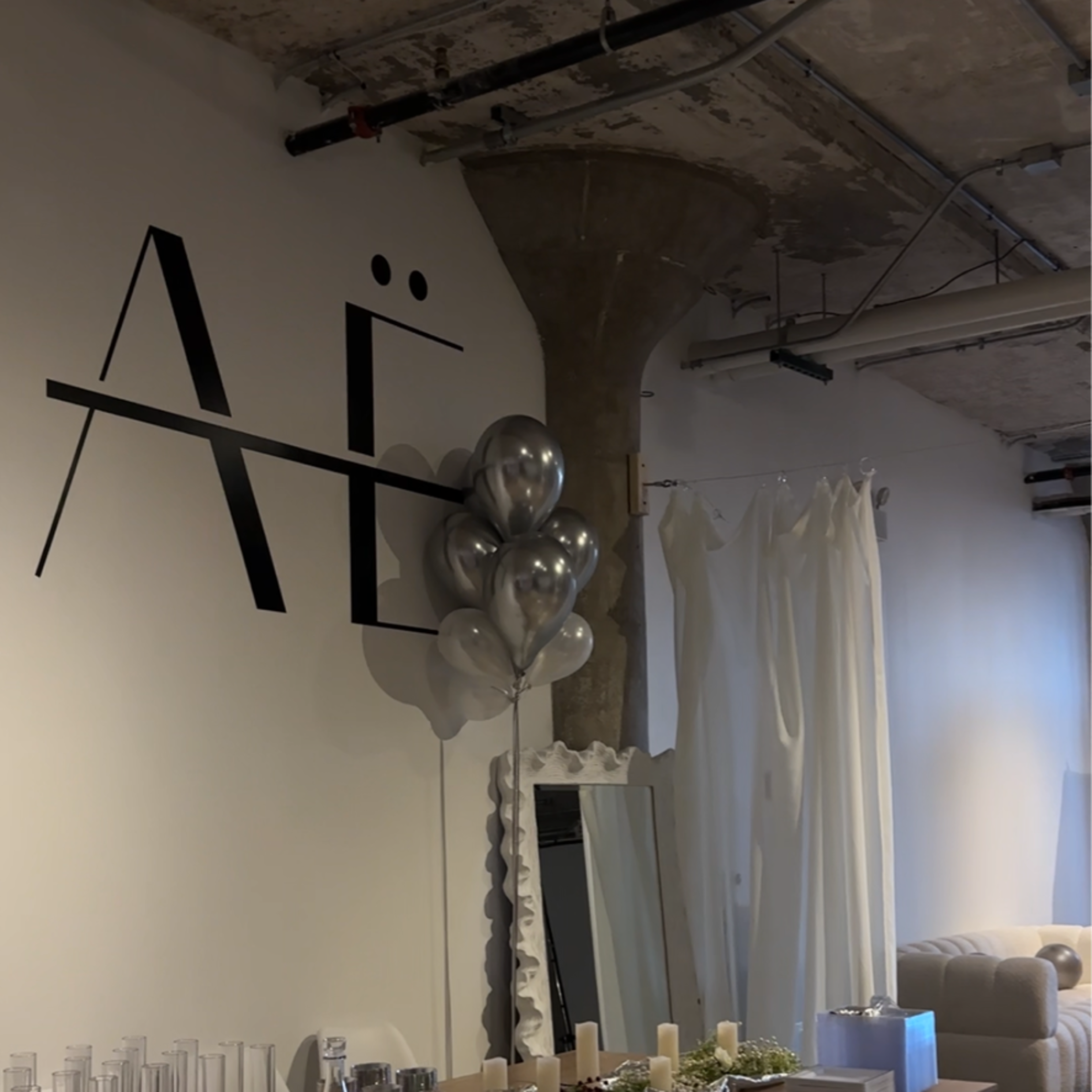 A modern event space with a long wooden table set for a gathering, adorned with champagne glasses, greenery, and silver balloons, against a backdrop of a minimalist white wall featuring large stylized letters.