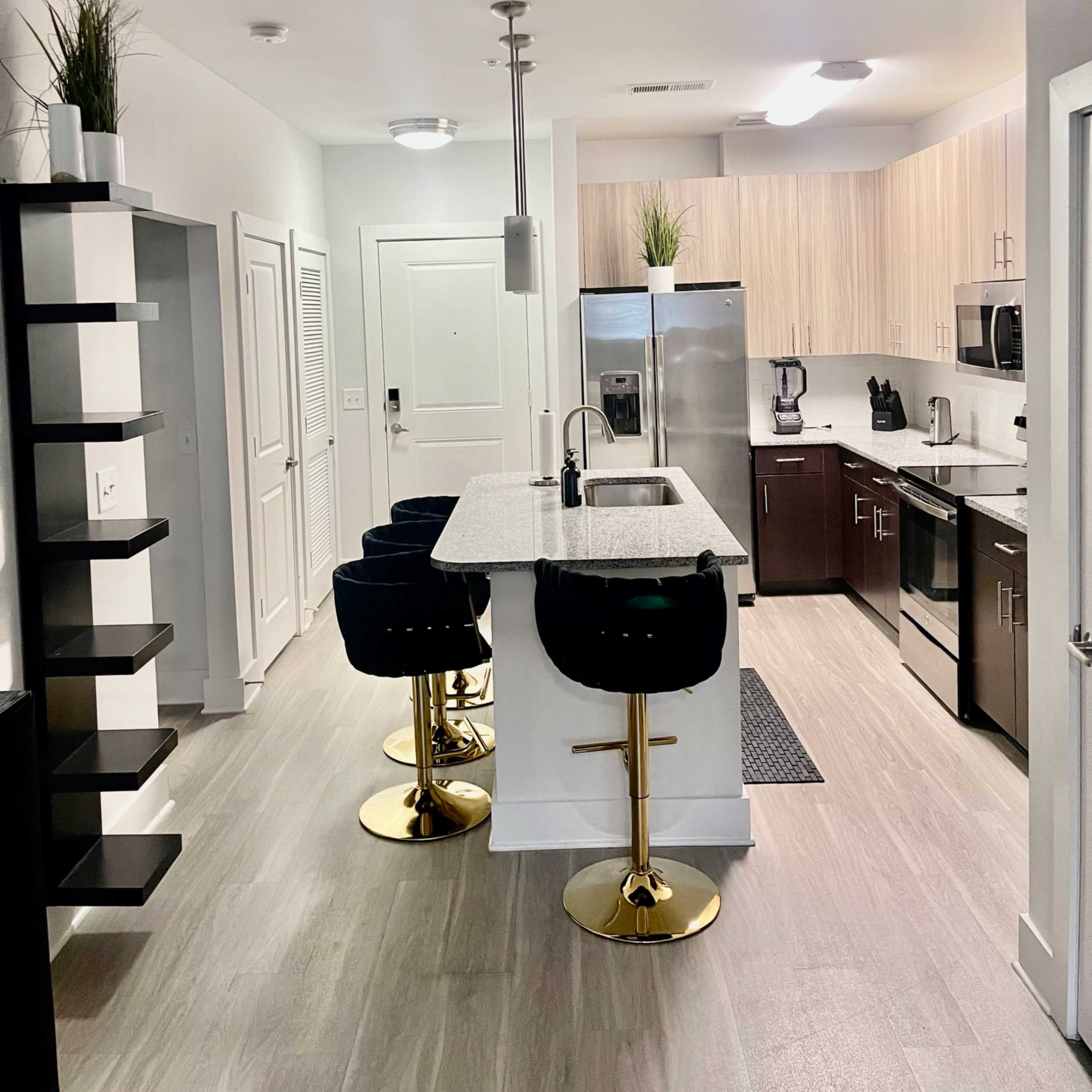 A modern kitchen featuring a central island with three black bar stools, stainless steel appliances, and light wood cabinetry.