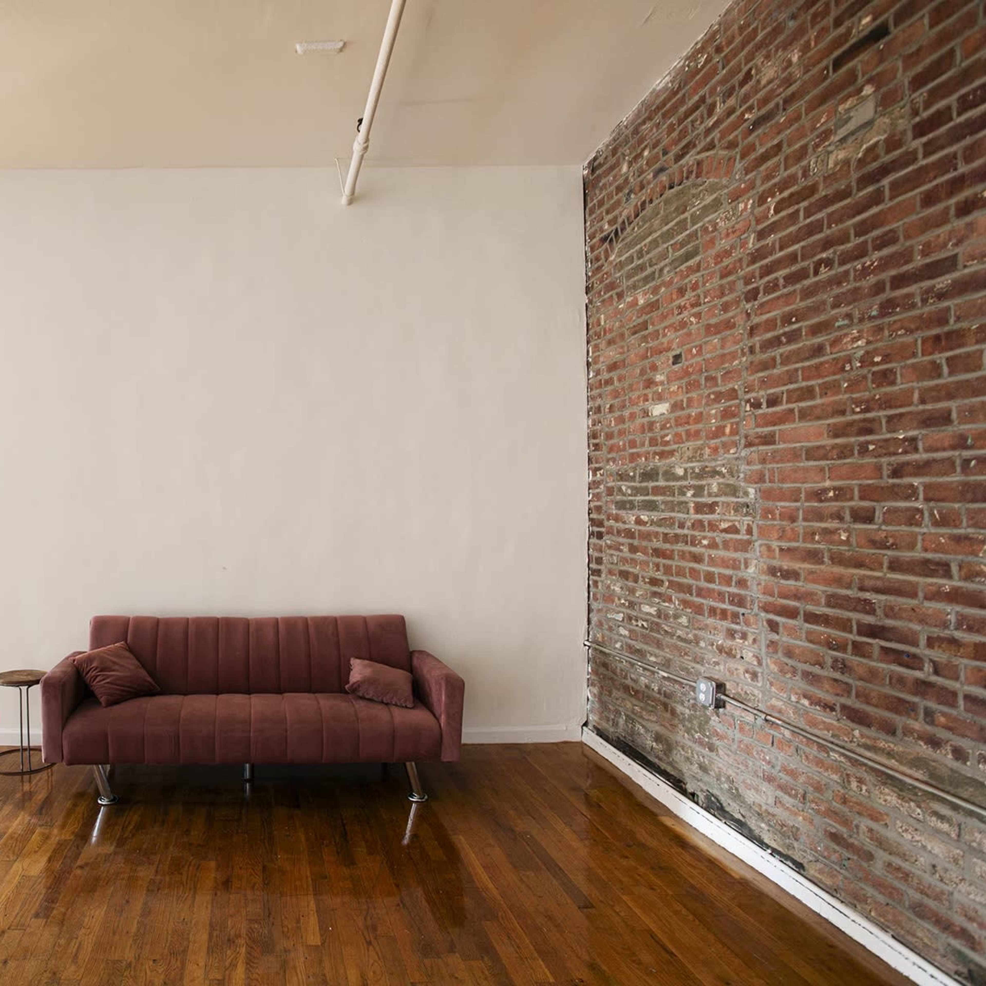 A minimalist interior features a brown sofa against a white wall, with exposed brick and wooden flooring.