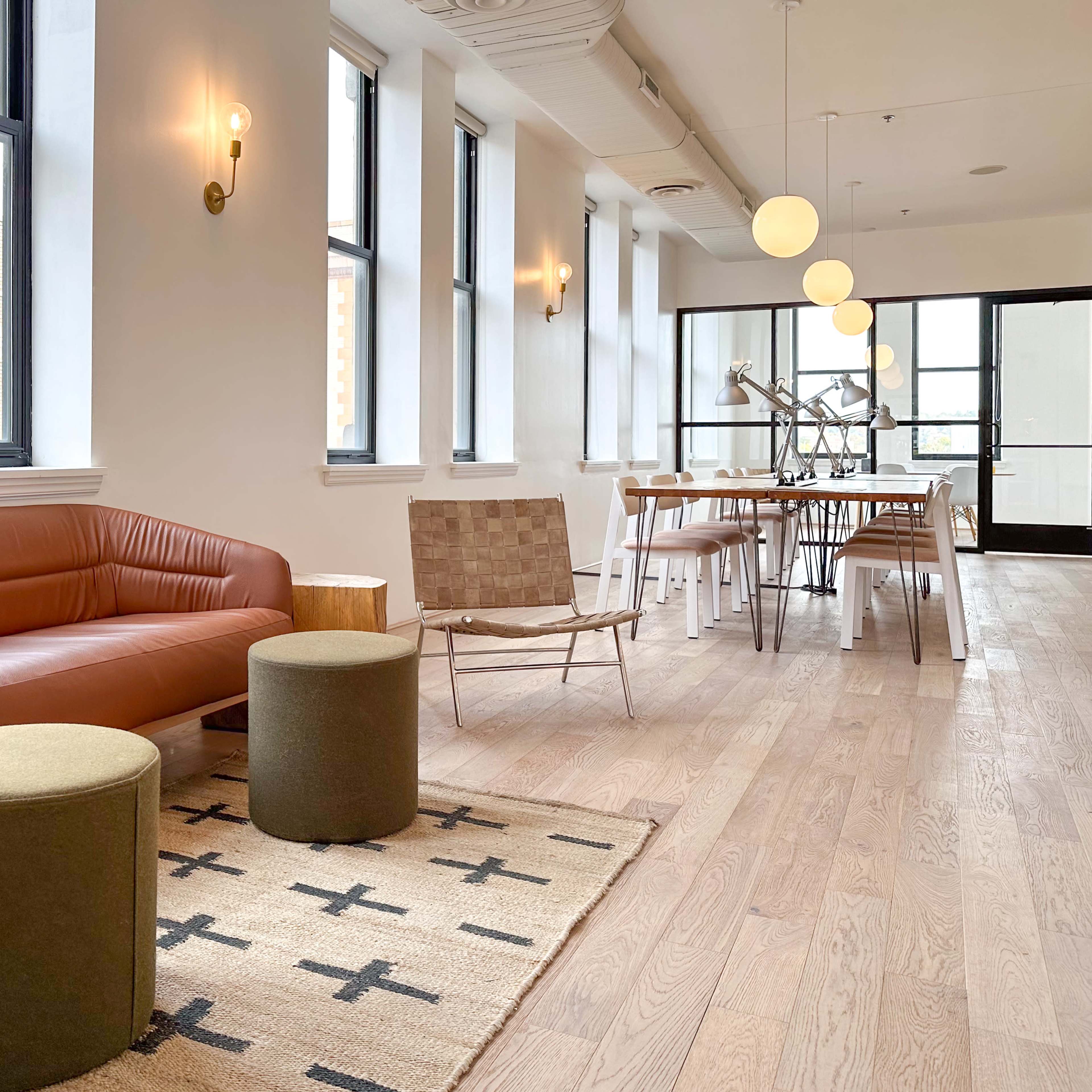 A modern, minimalistic interior featuring a brown leather sofa, two round stools, a woven area rug, a dining table with chairs, and large windows.