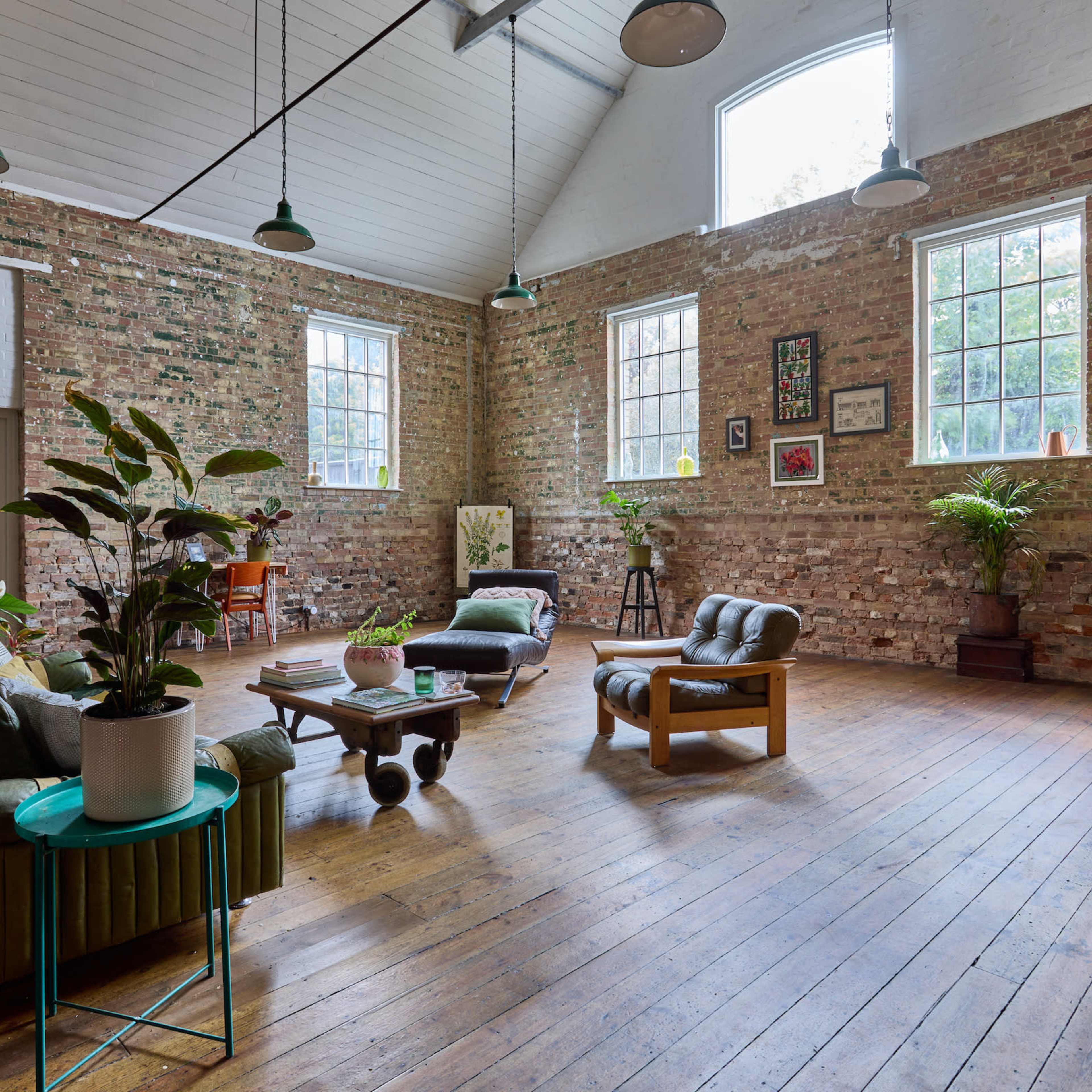 A spacious room with exposed brick walls, wooden floors, and large windows, furnished with green sofas, a wooden coffee table, and various plants.