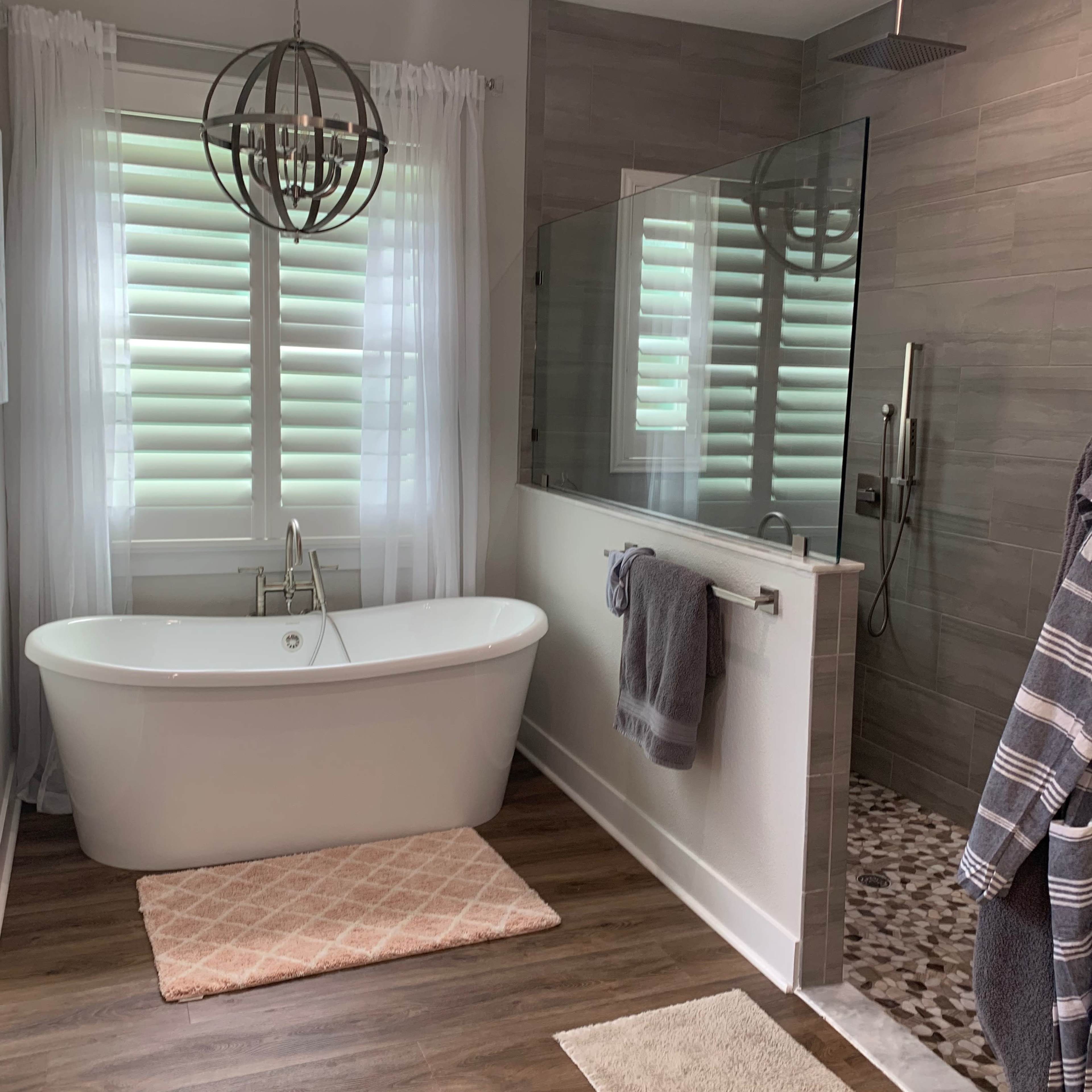 A modern bathroom featuring a freestanding bathtub, a glass shower enclosure, and natural light streaming through shuttered windows.