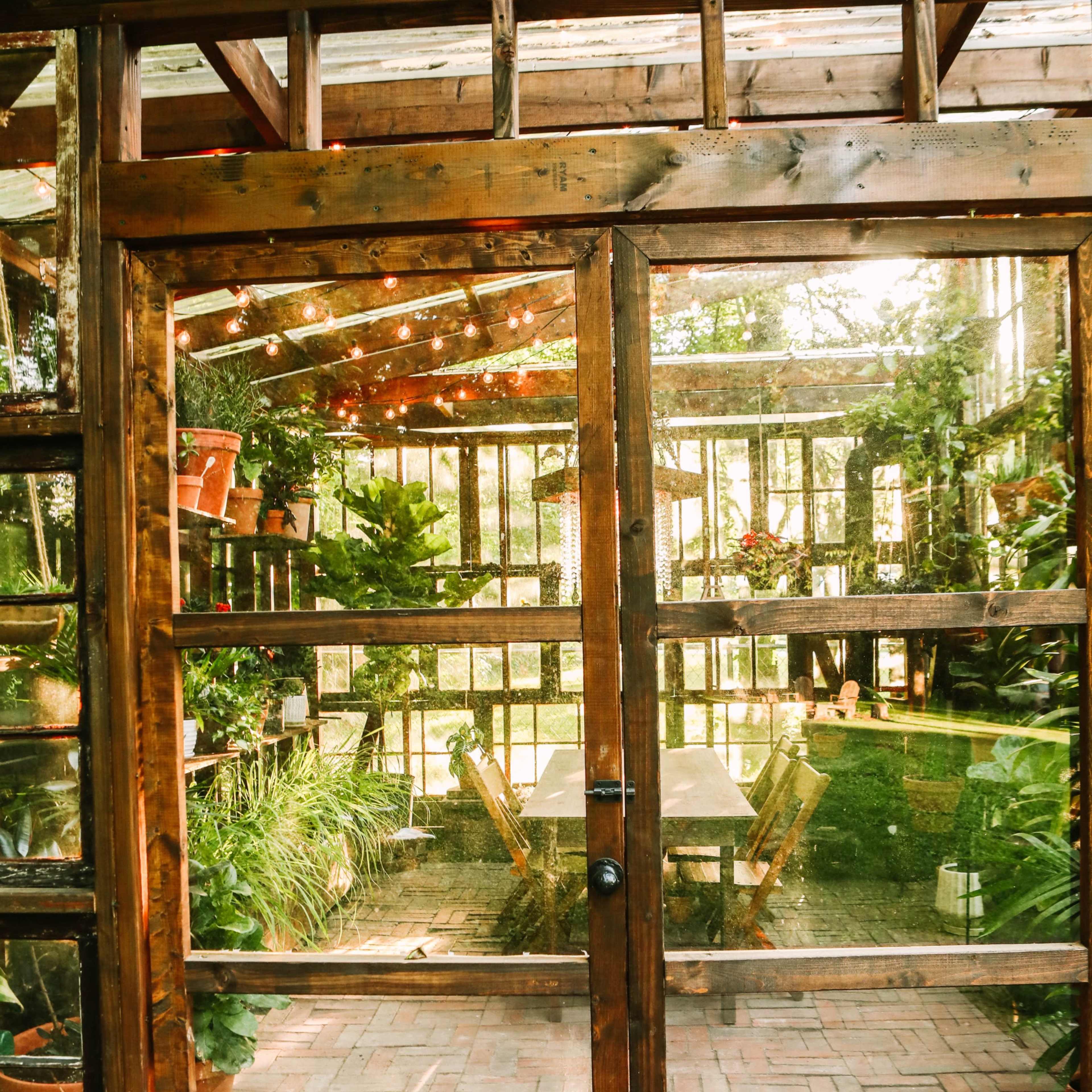 A wooden-framed greenhouse with a glass roof, filled with various plants and a set of tables inside, and a door that opens to the garden.