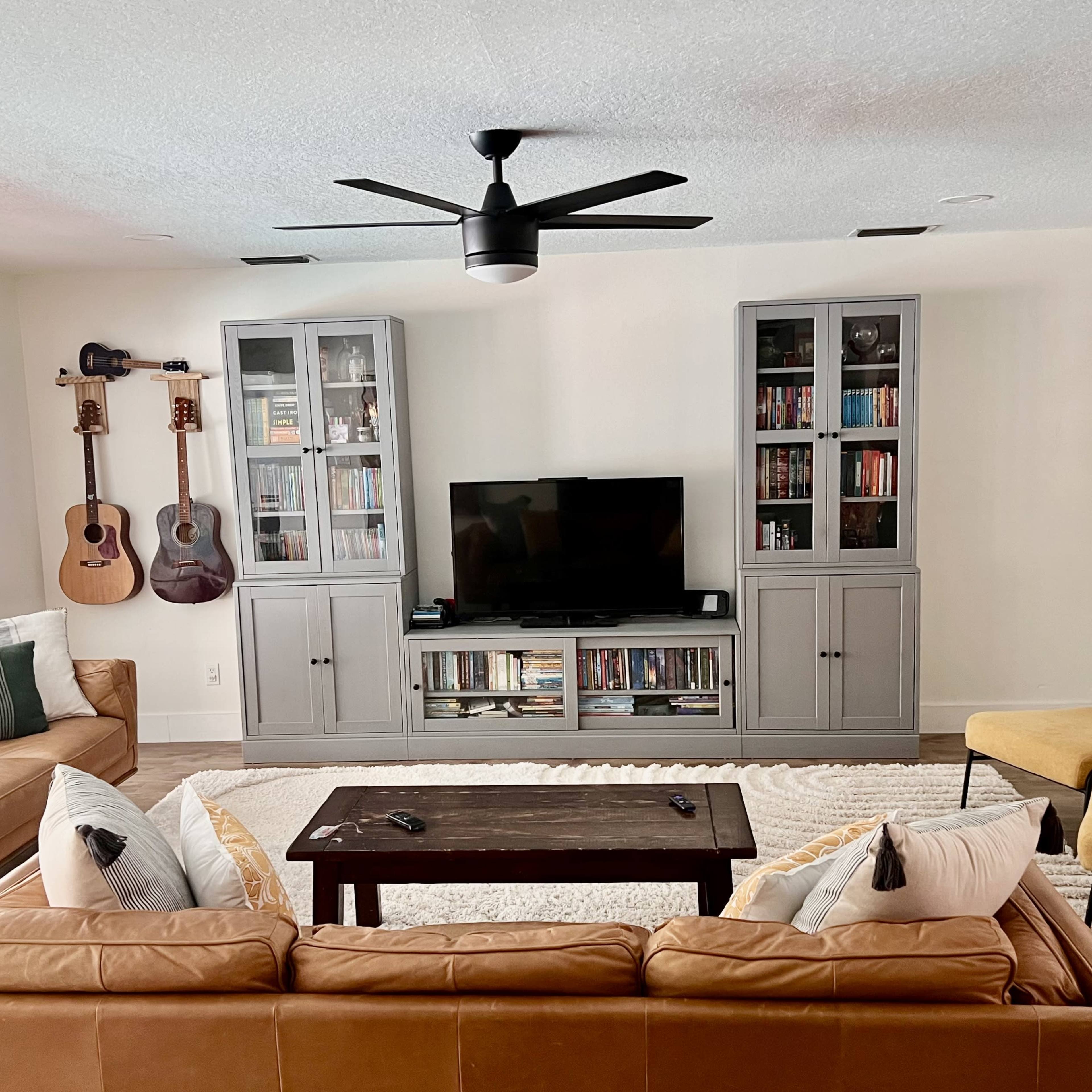 The living room features a brown leather sofa, two yellow chairs, a central coffee table, and a TV unit with bookshelves, surrounded by guitars on the wall and a light rug covering the floor.