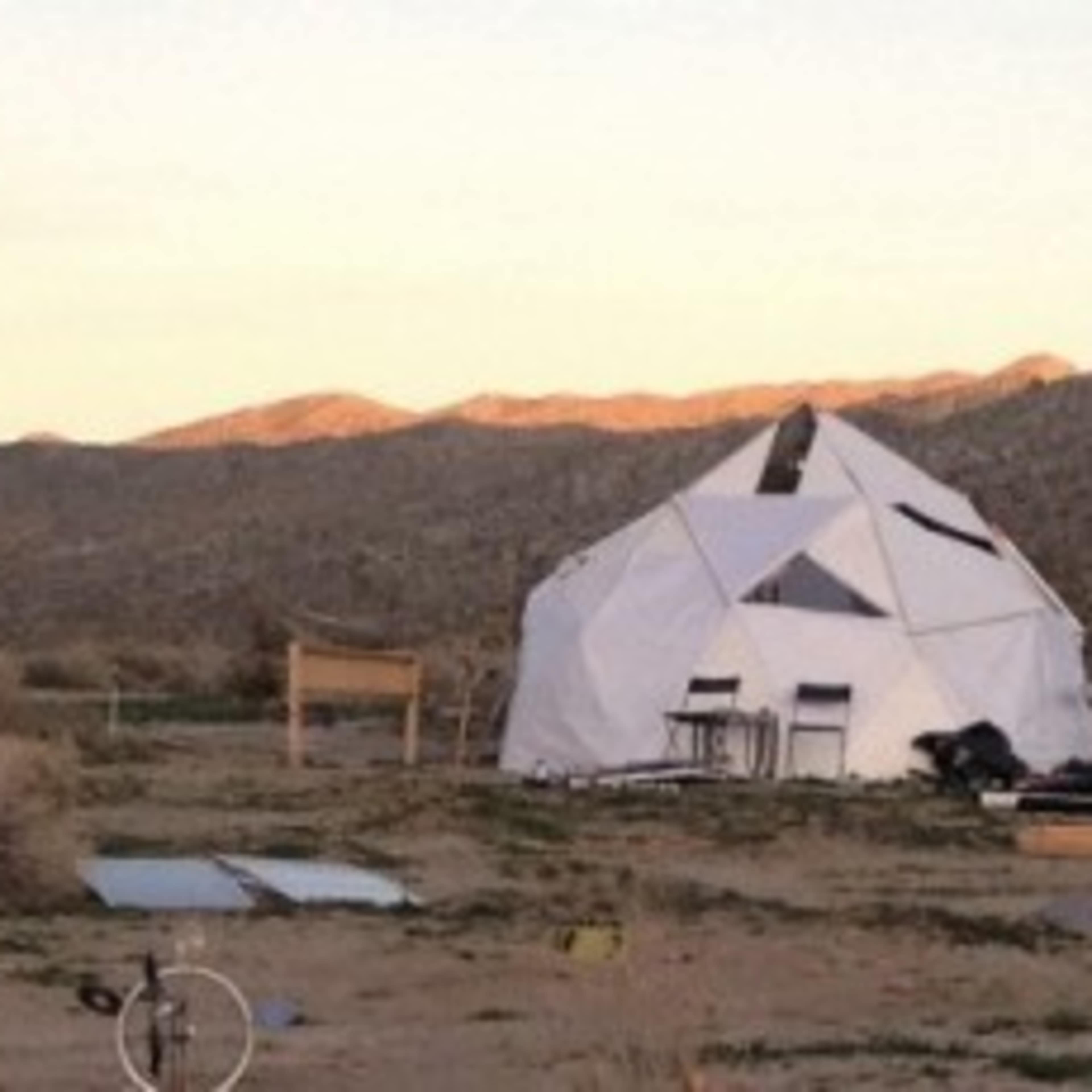 A temporary settlement with wooden structures and a geodesic dome is set against a backdrop of mountains at sunset.