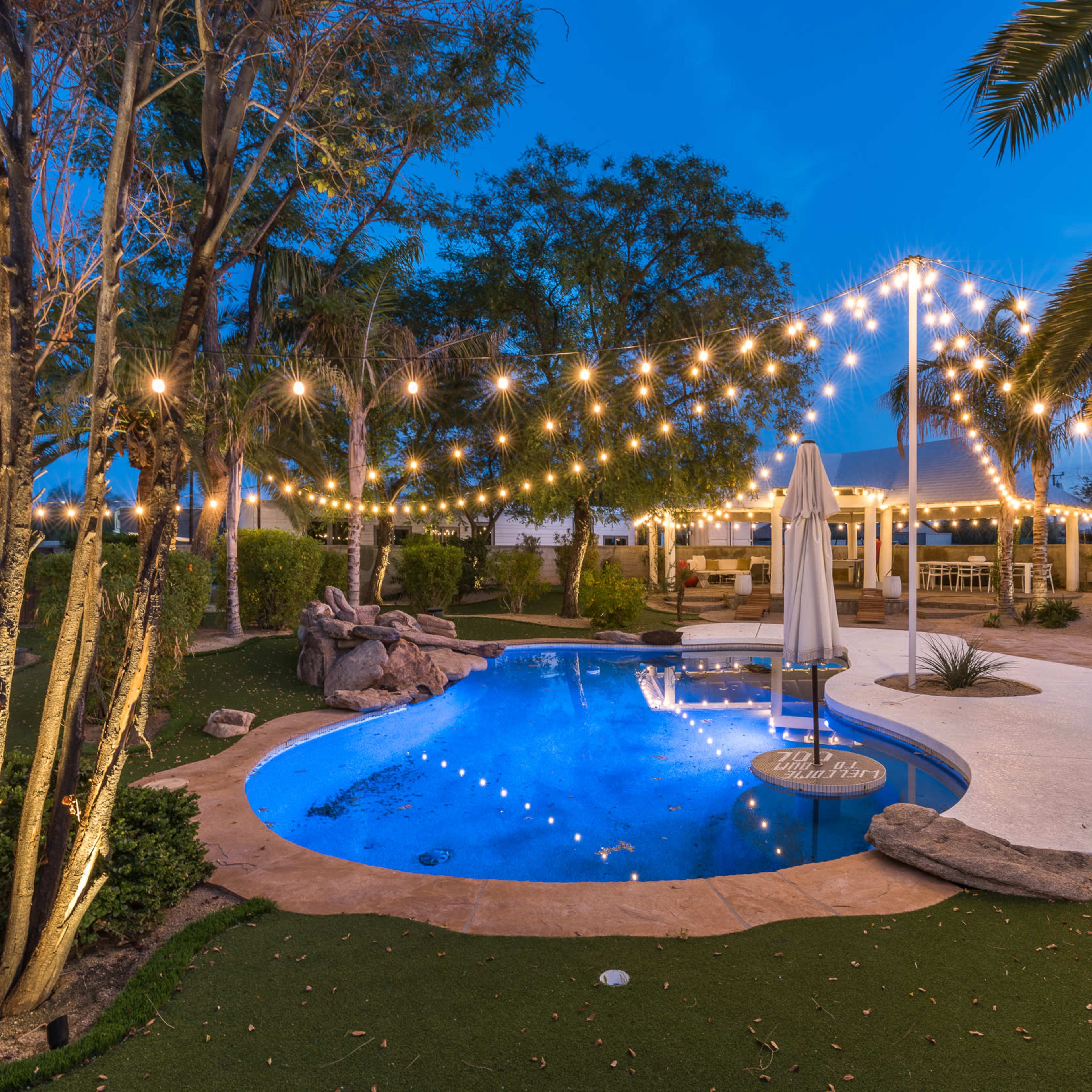 A backyard pool area with string lights above, surrounded by landscaped greenery and a shaded patio.