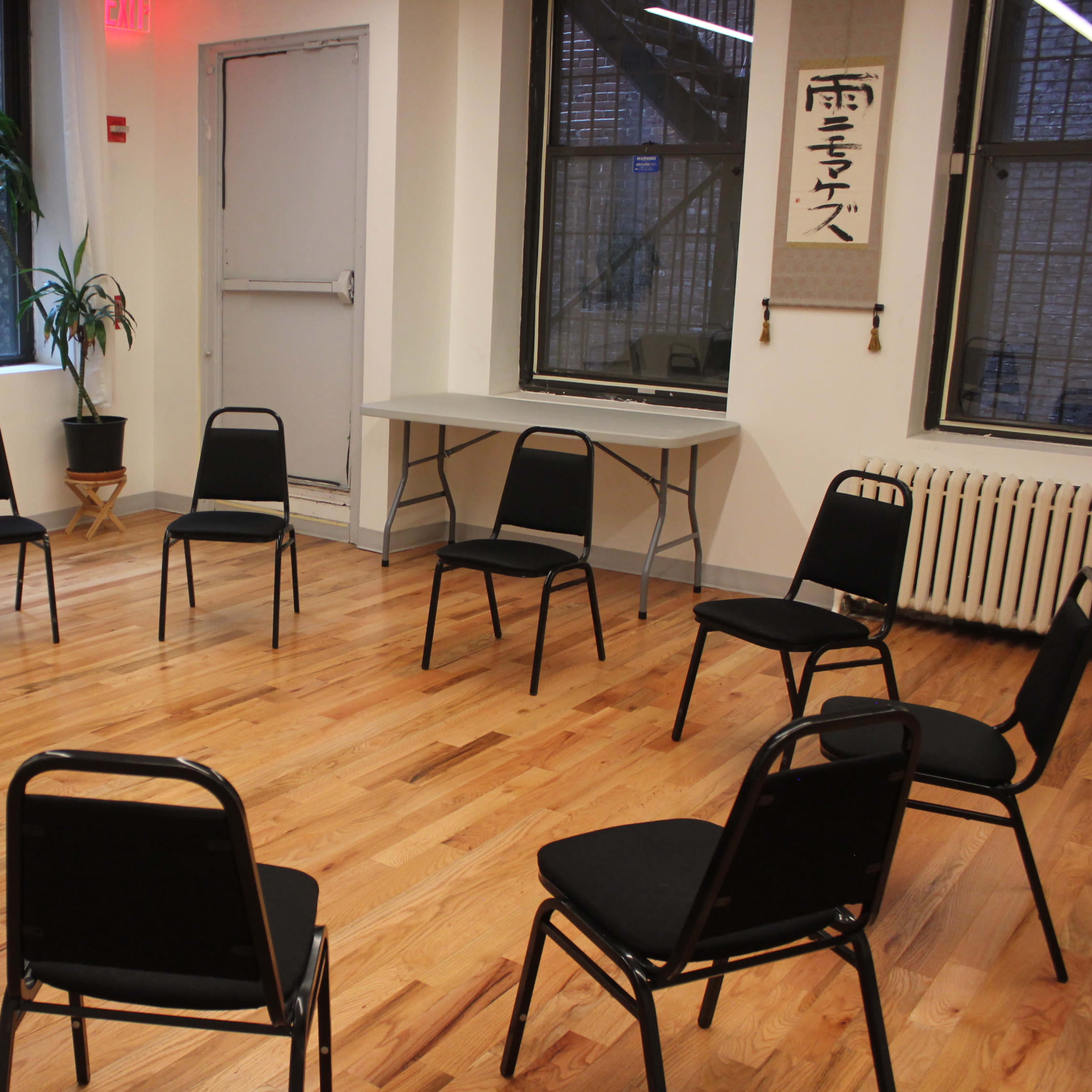 A circle of black chairs is arranged on a wooden floor in a well-lit room with large windows and a potted plant.