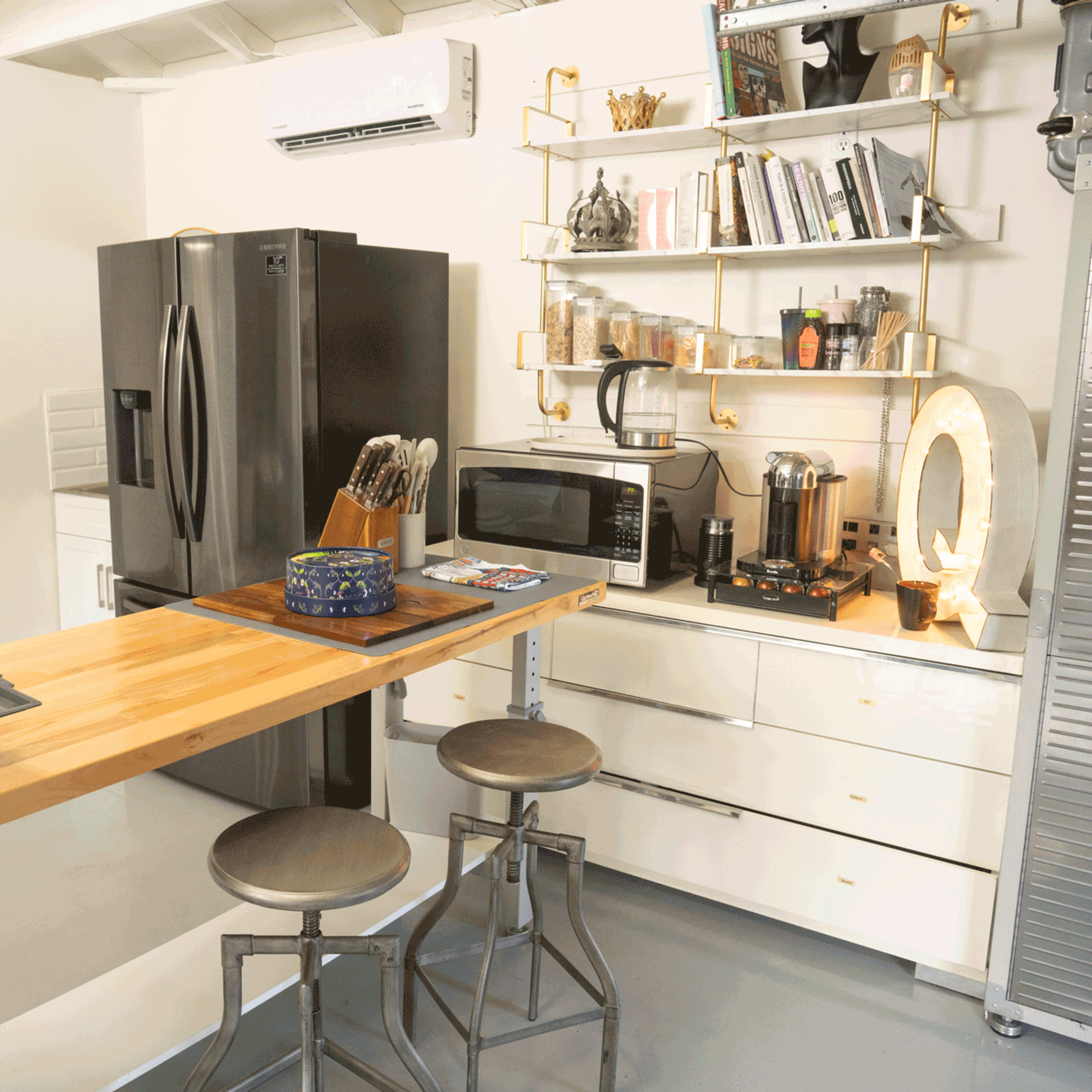 A modern kitchen with a wooden island and two metal stools, featuring a black refrigerator, microwave, coffee maker, and shelves filled with books and kitchen items.