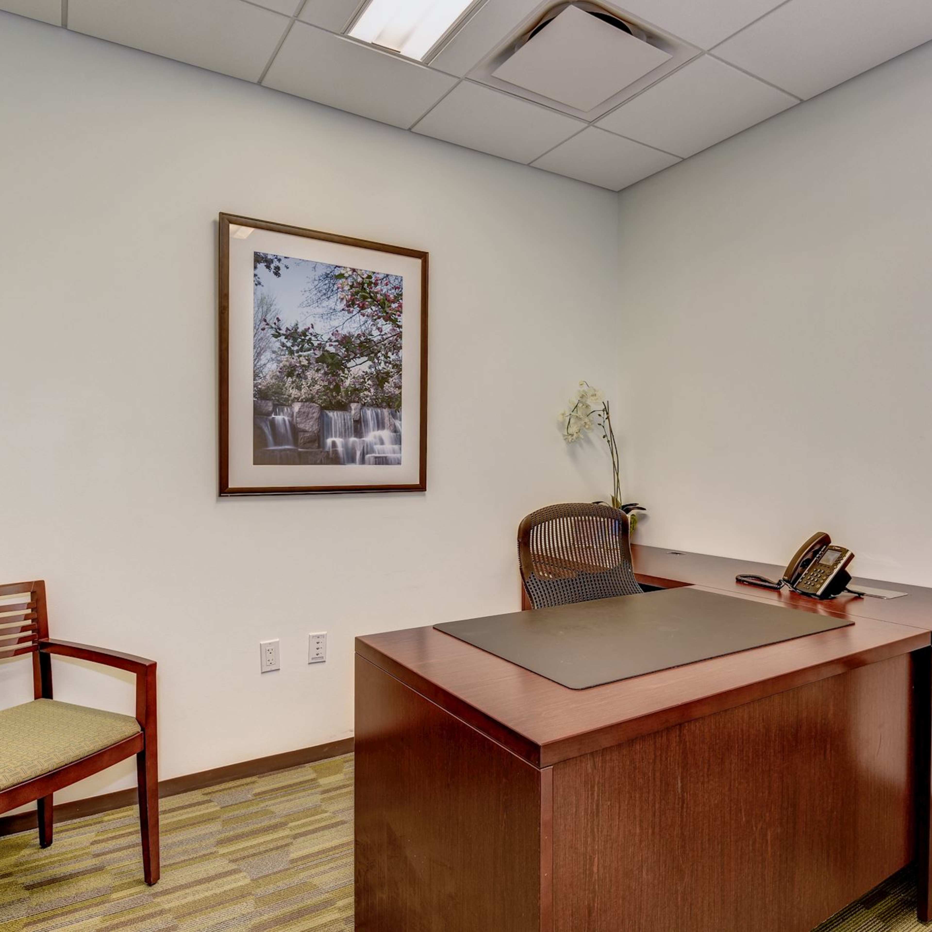 A simple office space with a wooden desk, an ergonomic chair, two wooden chairs, and a wall-mounted framed picture.