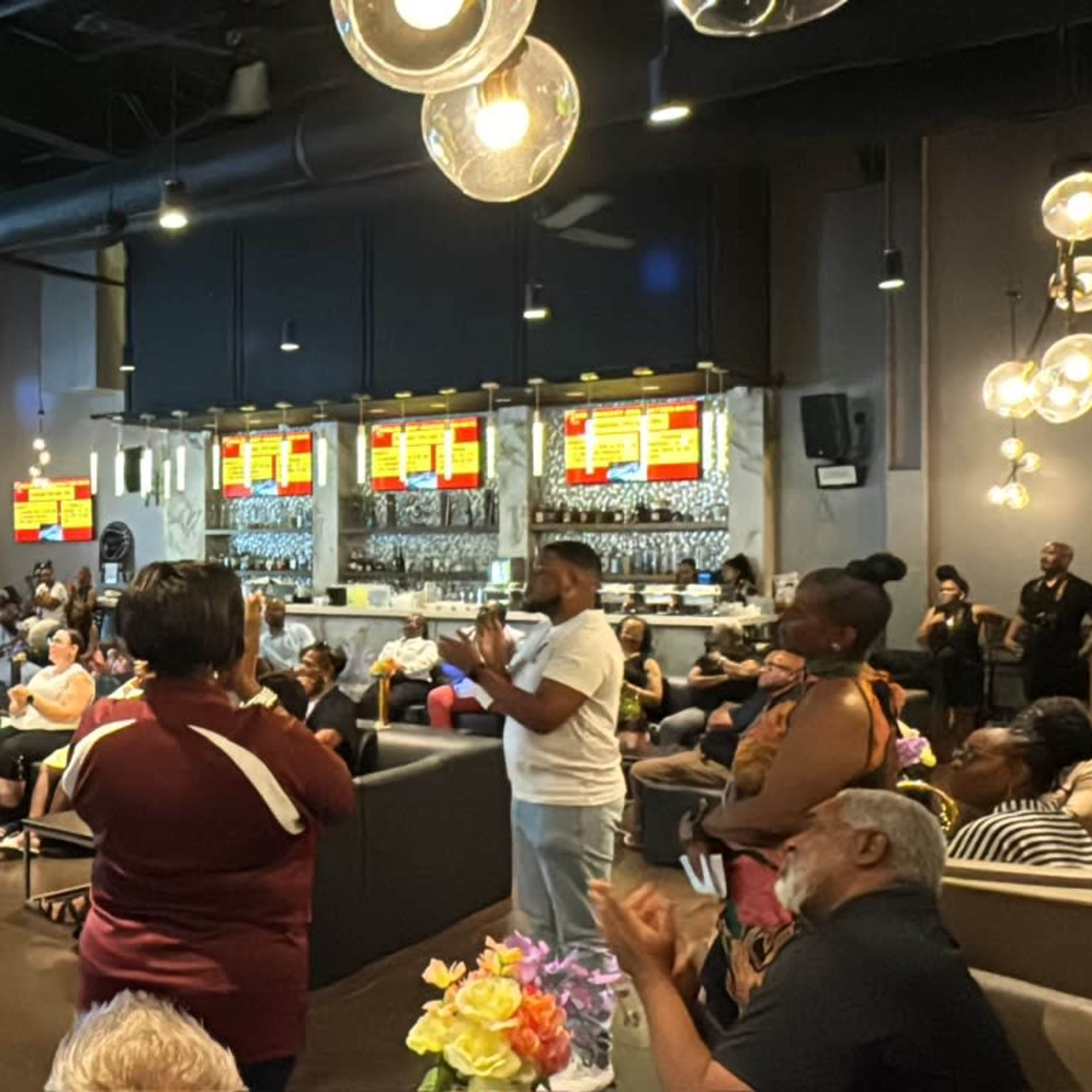 A speaker stands in front of a large audience in a modern event space with a bar in the background.