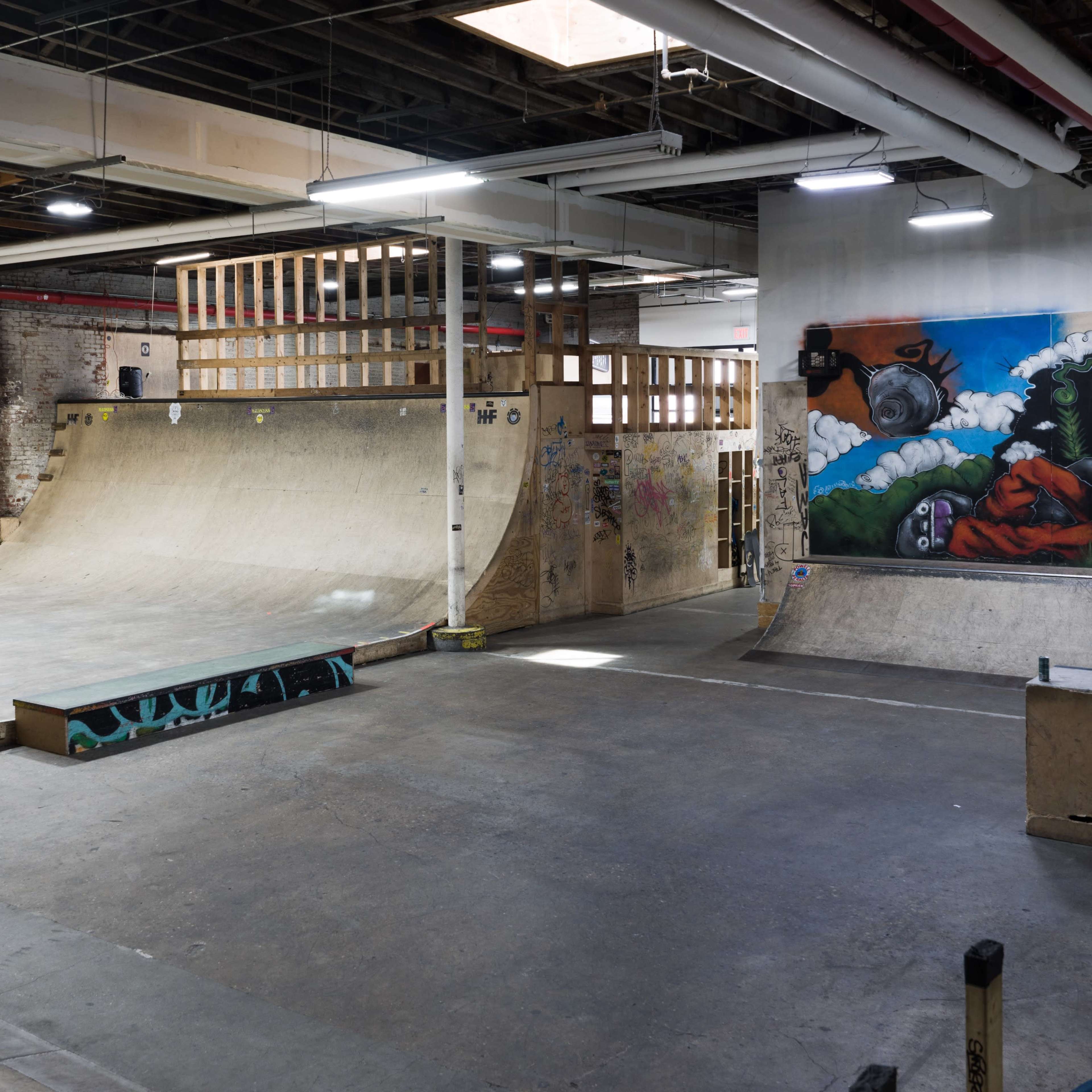 An indoor skatepark with a wood ramp, a half-pipe, and a mural on the wall.