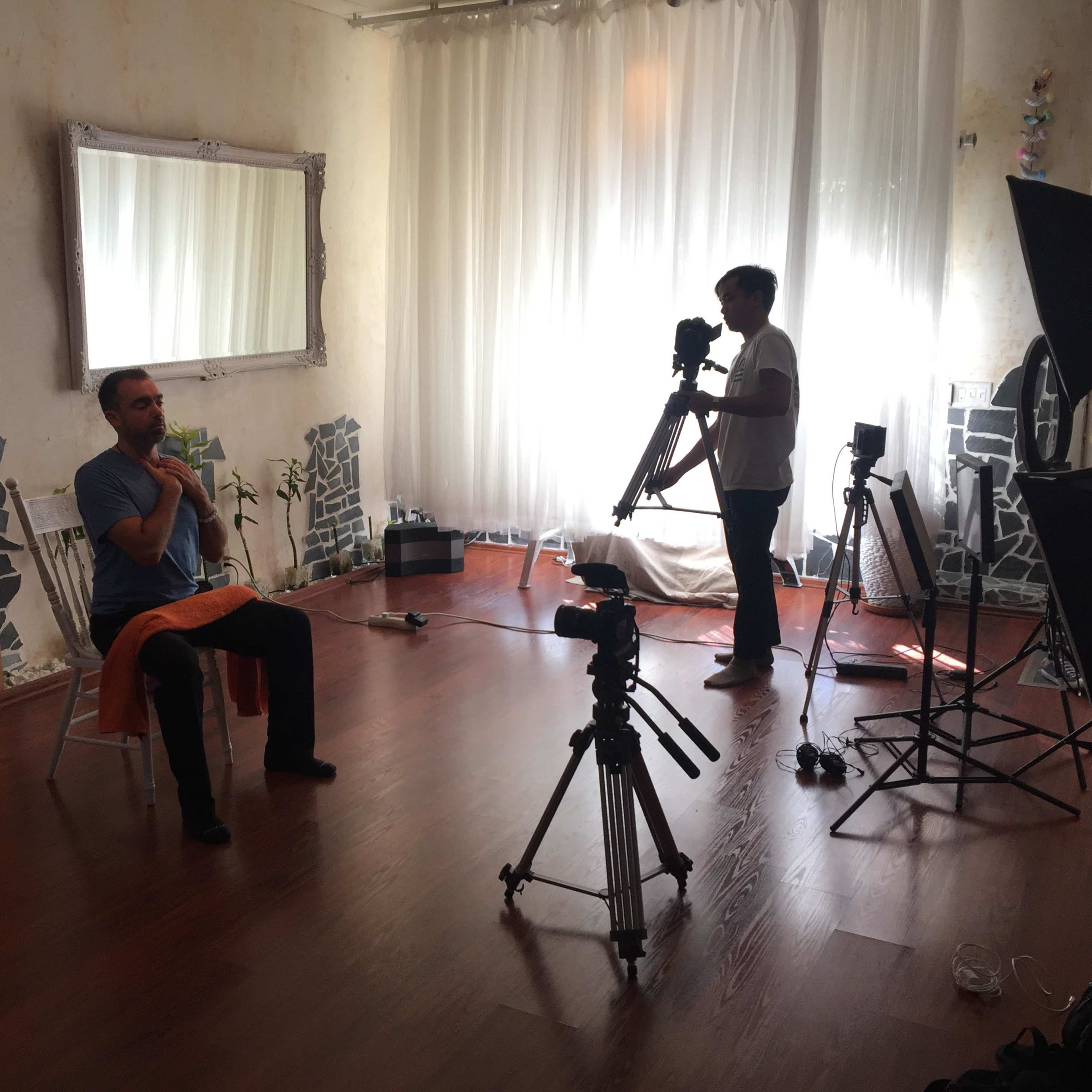 A person sits on a chair in a well-lit room while another person adjusts a camera on a tripod.