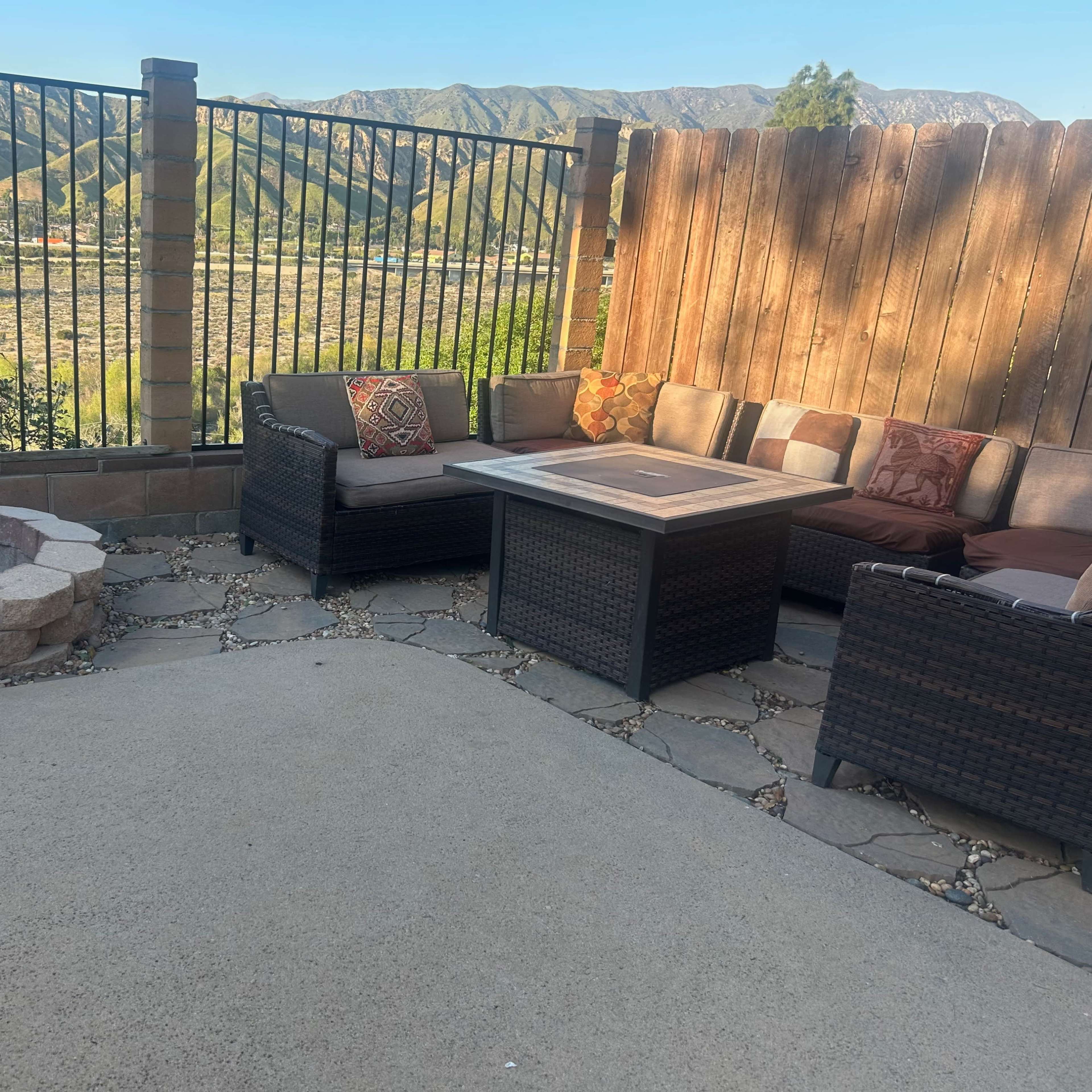 A patio area features a stone fire pit, a table, and wicker seating surrounded by a fence with mountains in the background.