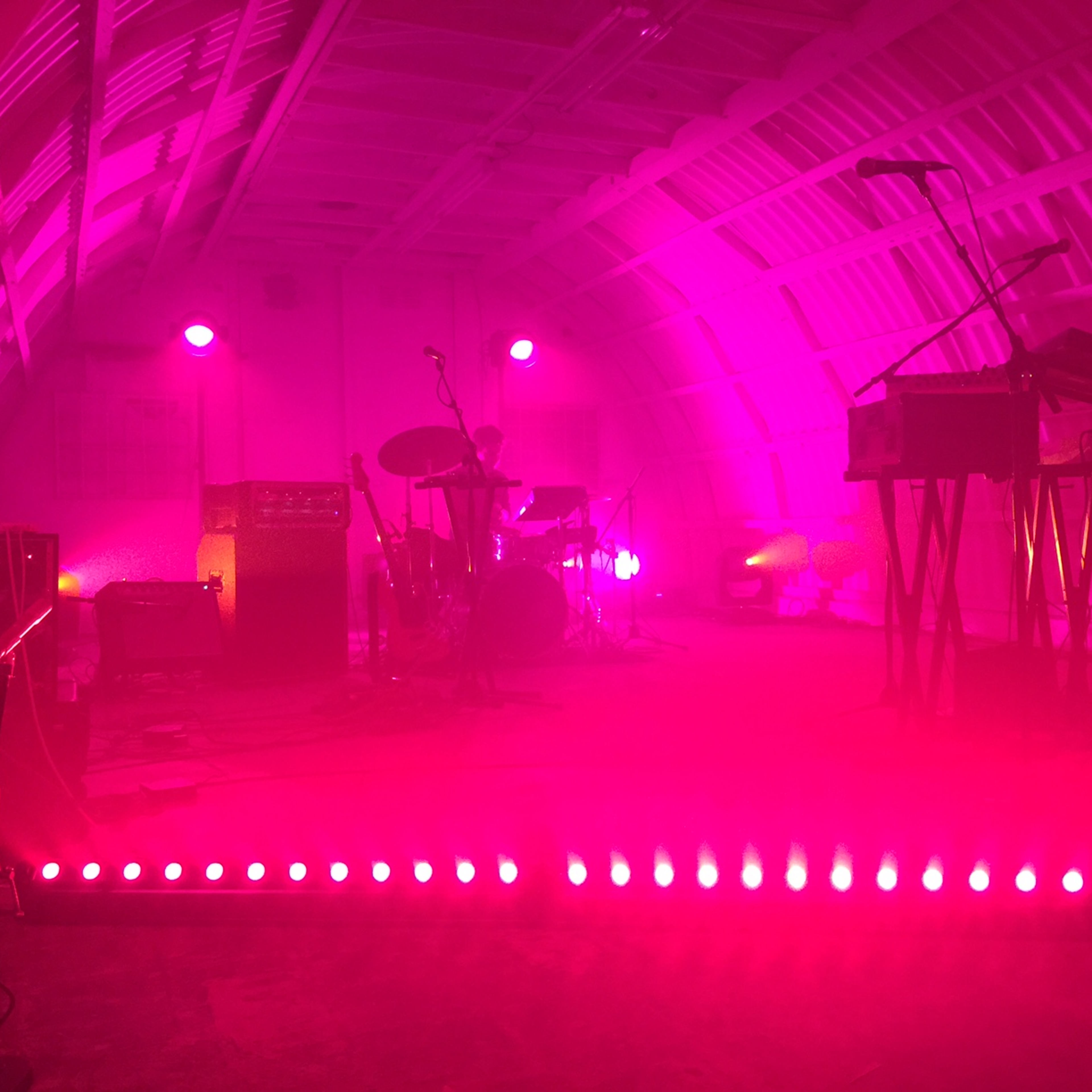 A music stage setup inside a brightly lit, pink-tinged venue with various instruments and equipment positioned for performance.