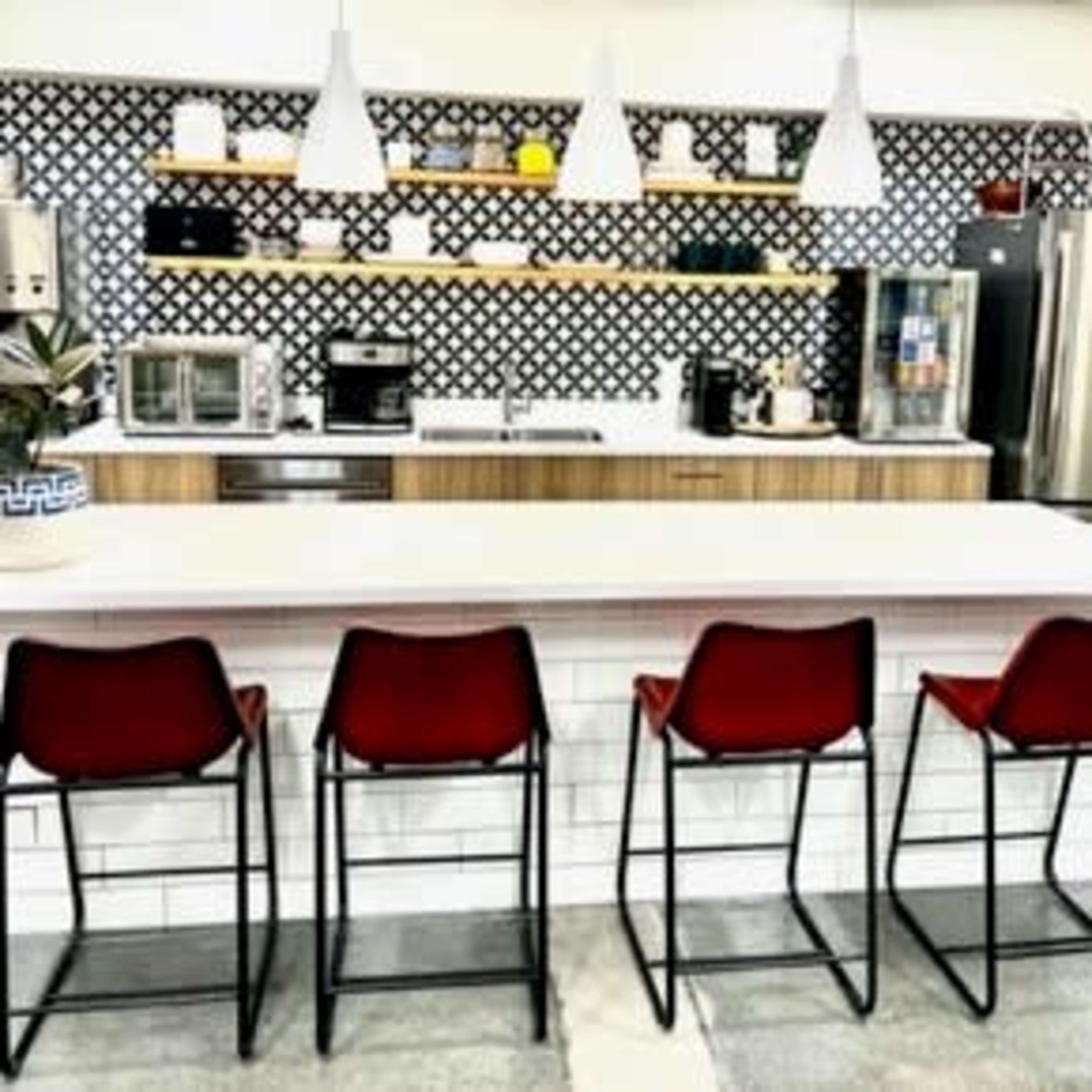 A modern kitchen features a long countertop with four red stools, a patterned backsplash, and various appliances on wooden shelves.