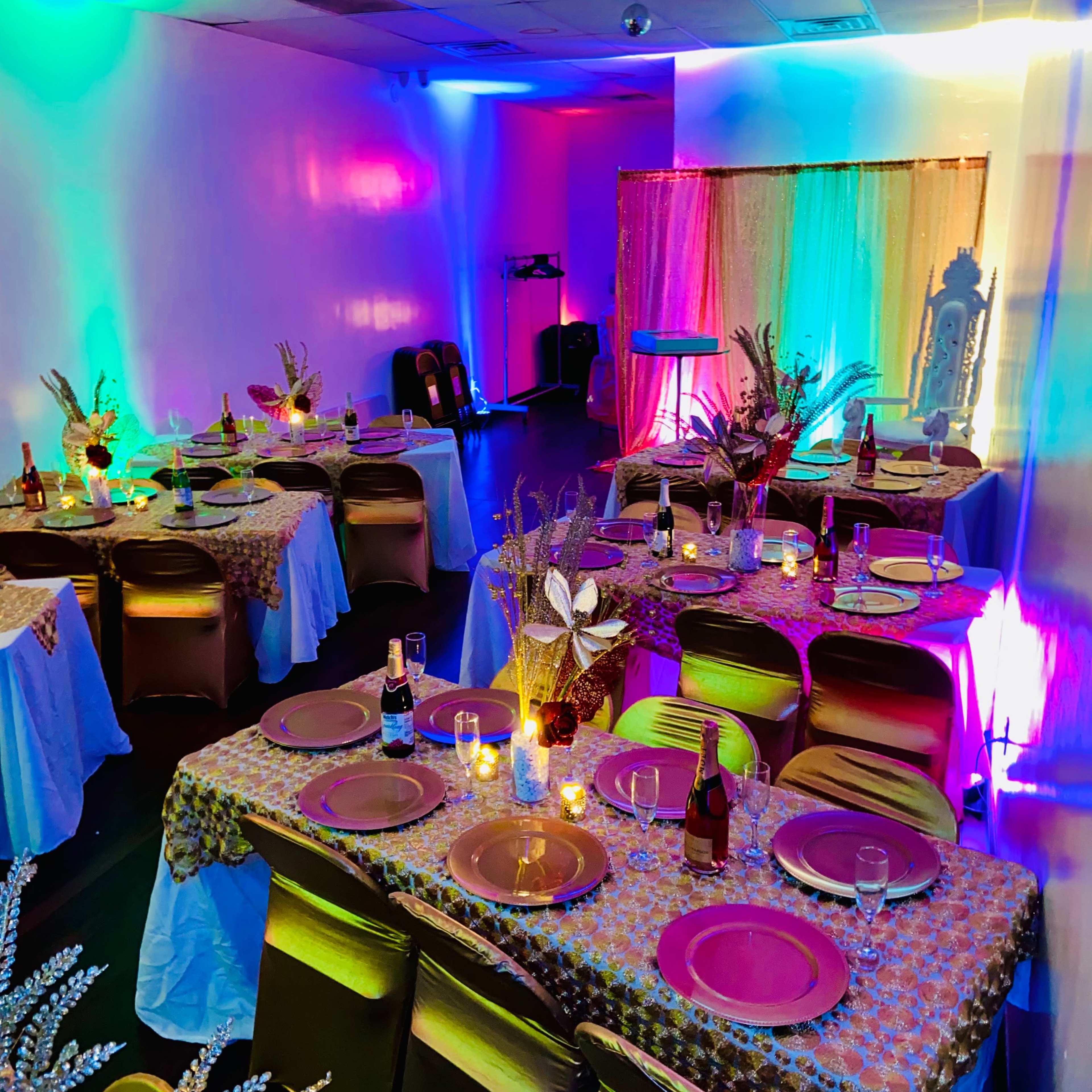 A banquet room with tables set for a celebration, featuring colorful lighting and decorative centerpieces.