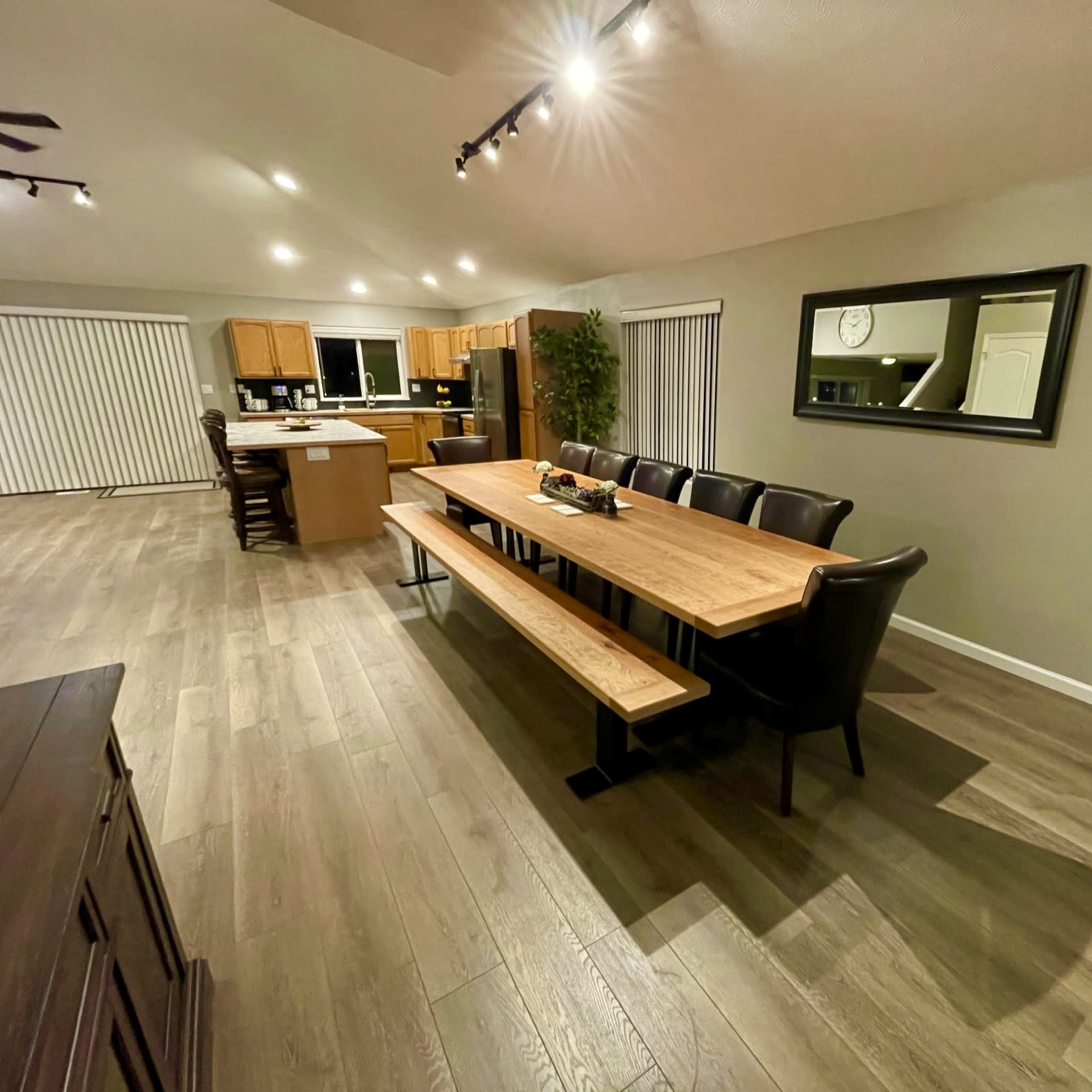 A spacious dining area with a long wooden table surrounded by black chairs, adjacent to a kitchen with light-colored cabinets.