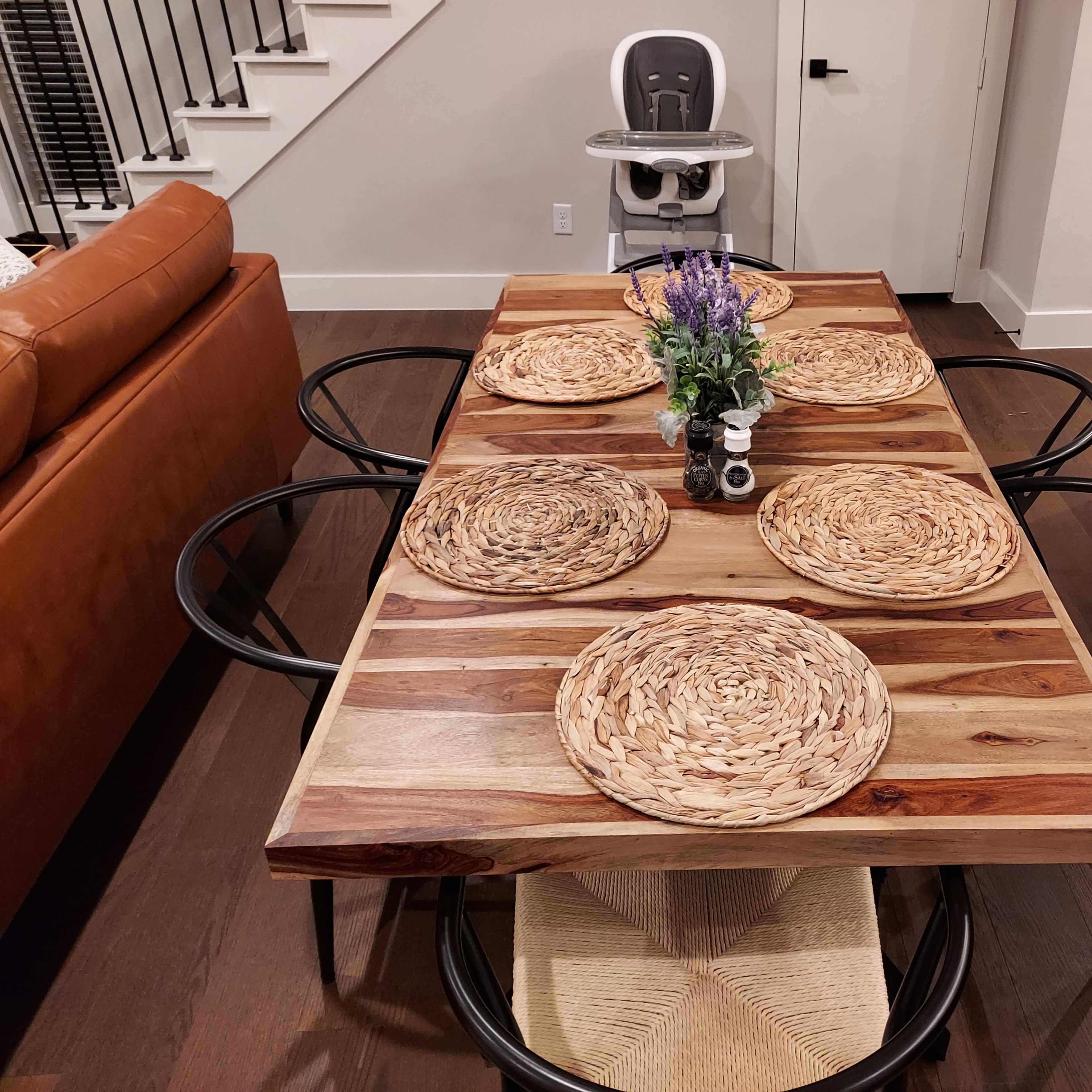 A wooden dining table set for six with woven placemats, surrounded by chairs, and a high chair visible in the background.