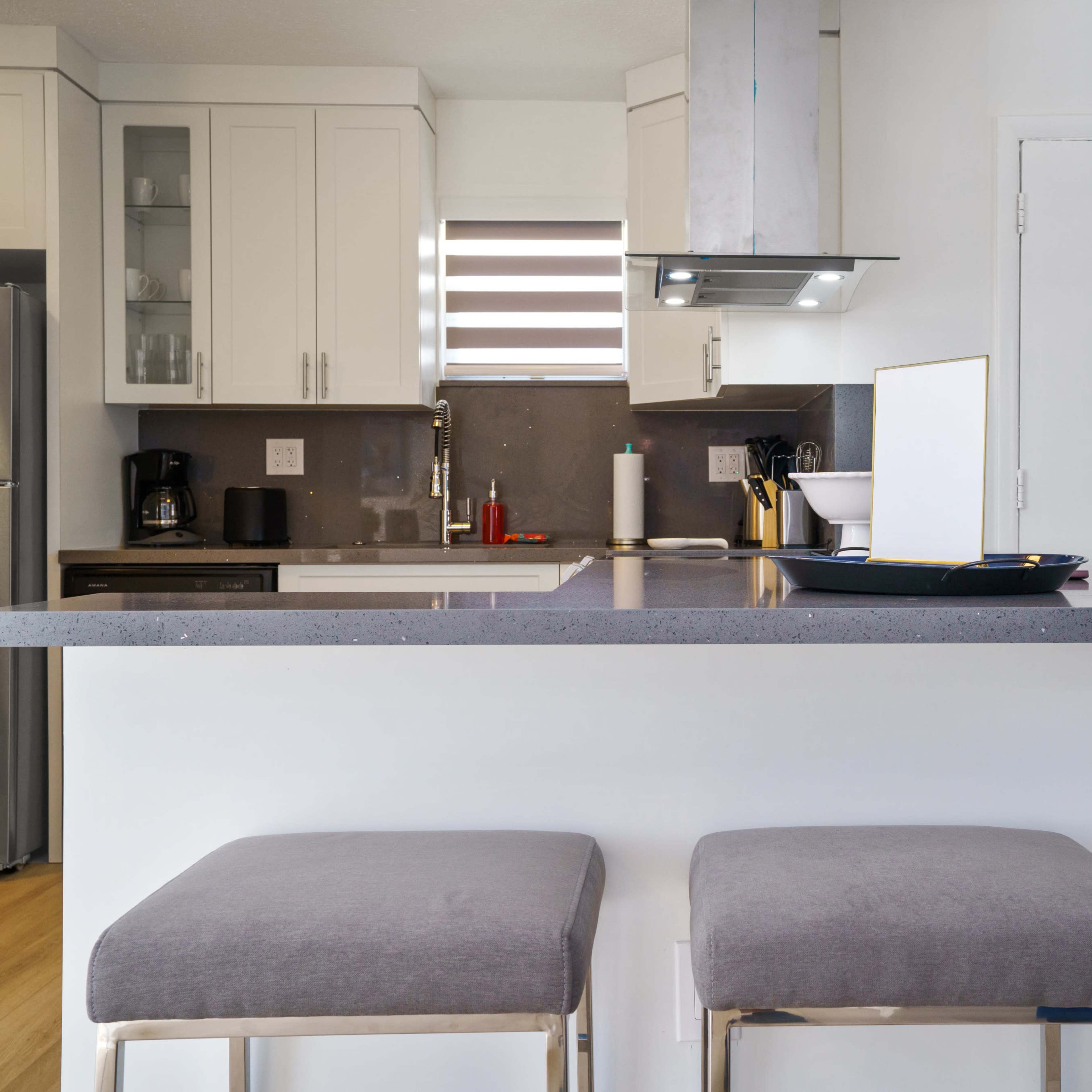The modern kitchen features white cabinetry, a gray countertop with two stools, stainless steel appliances, and a door leading to an exterior space.