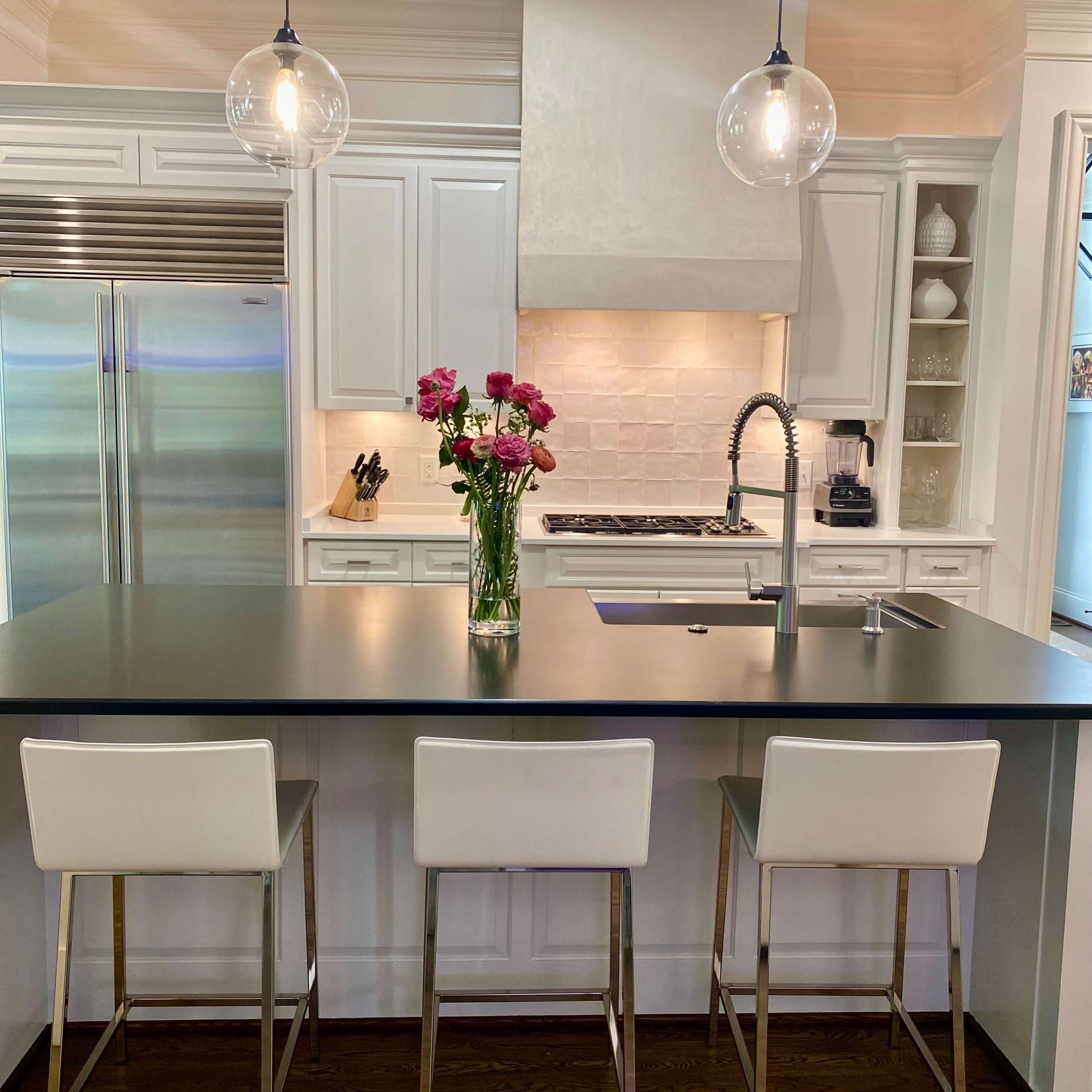 A modern kitchen features a central island with three white bar stools, a stainless steel sink, and a vase of pink flowers on the counter.