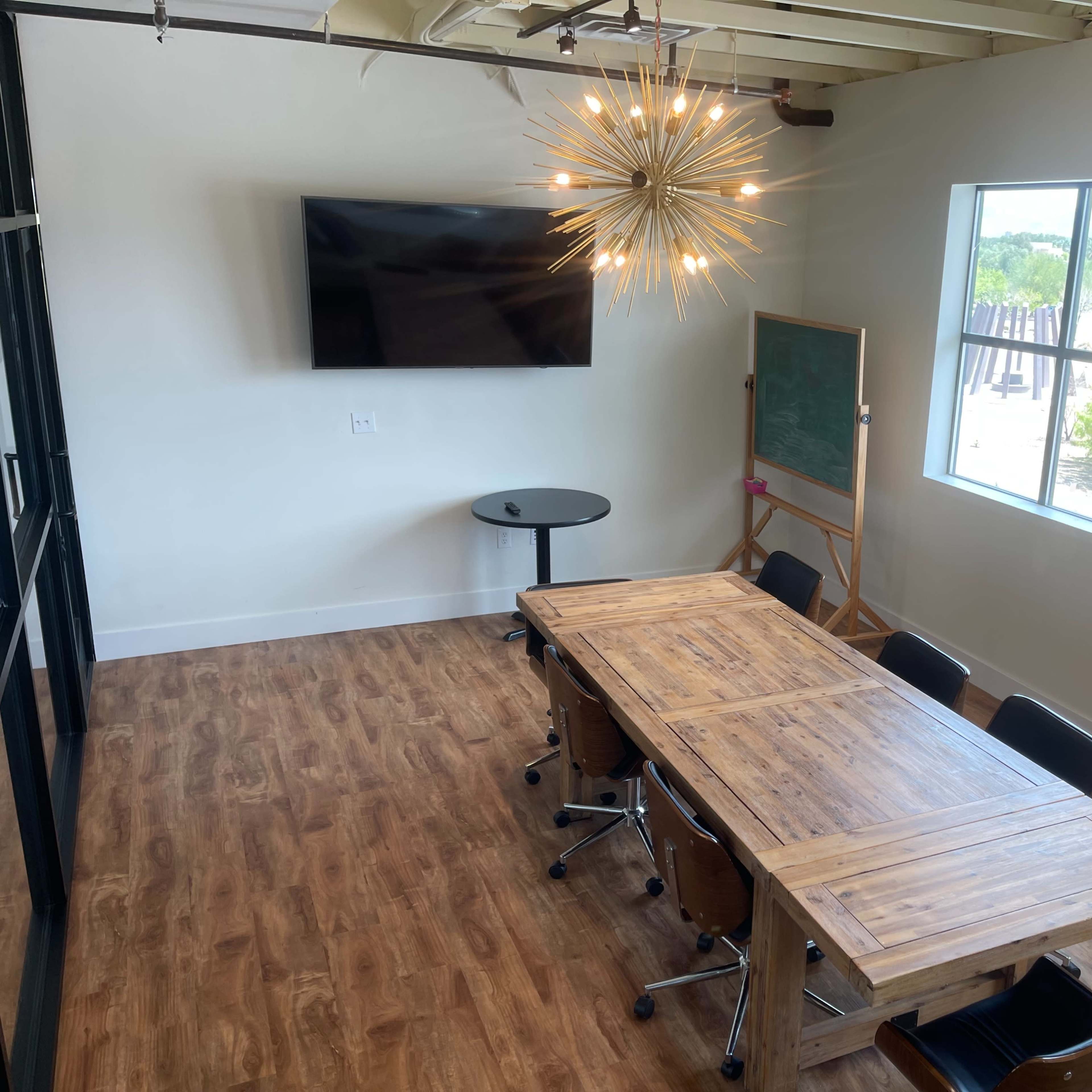 A modern conference room with a long wooden table, black chairs, a wall-mounted television, and a large window.