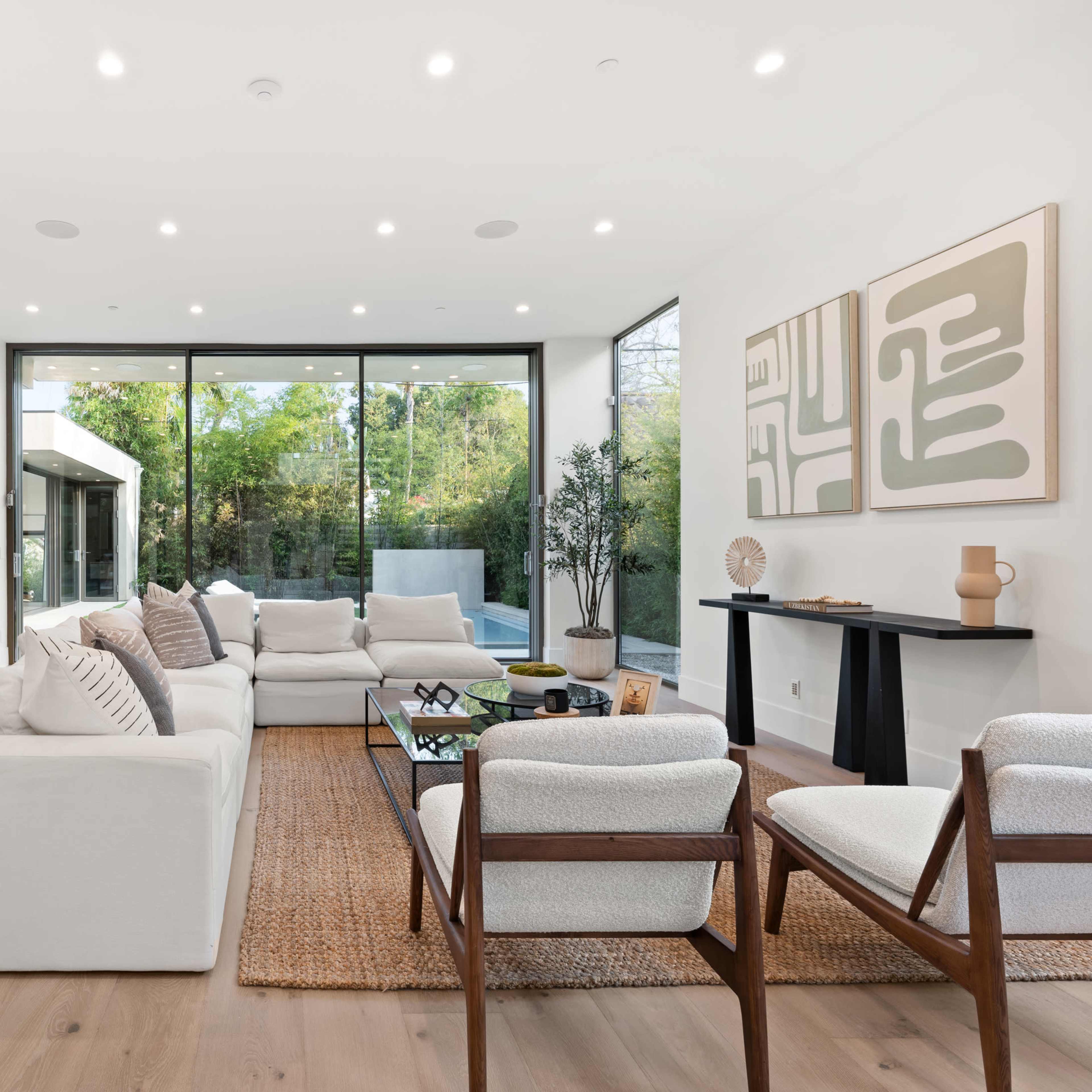 The modern living room features a large sectional sofa, minimalist furniture, and floor-to-ceiling windows that overlook a garden.
