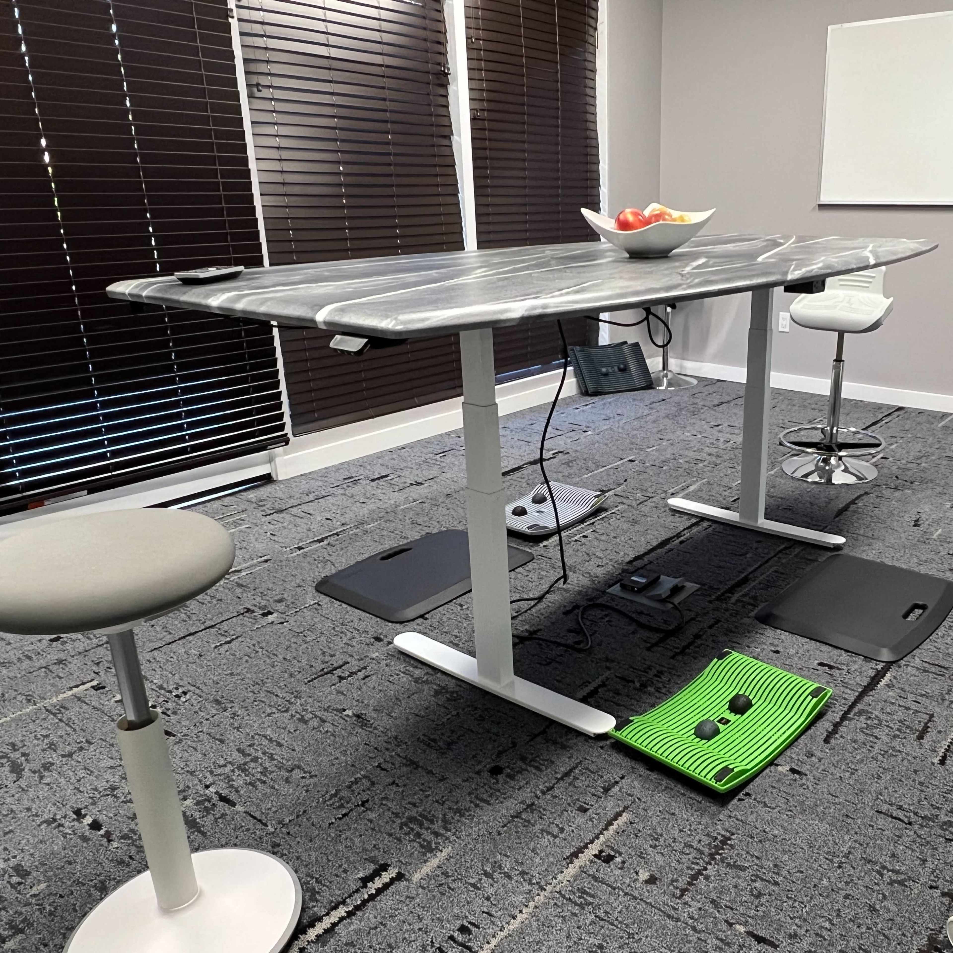 A modern meeting room features a height-adjustable table with a bowl of fruit on top, surrounded by ergonomic chairs and a green mat on the floor.