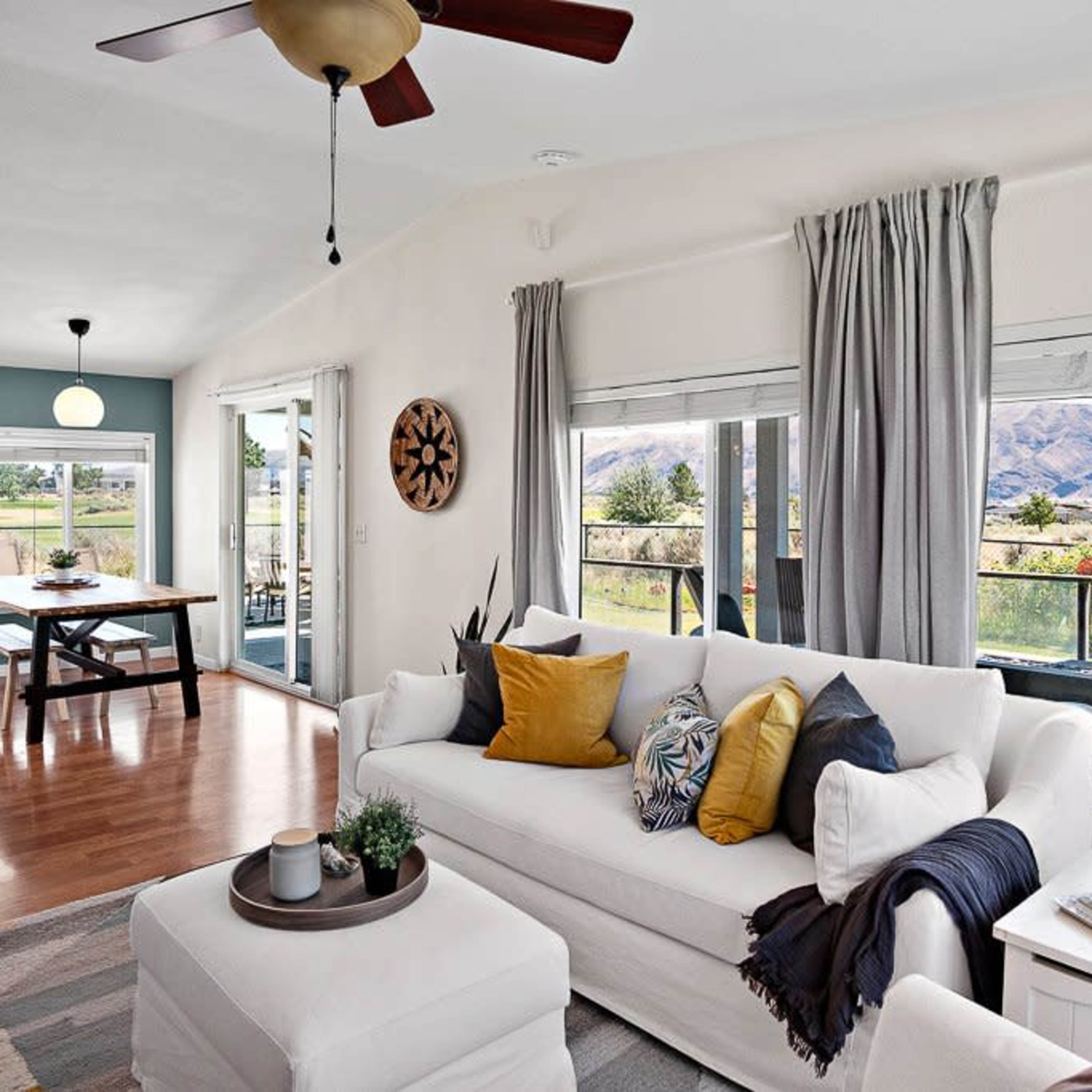 A bright living room features a white sofa adorned with decorative pillows, large windows with views of a natural landscape, and a dining area in the background.