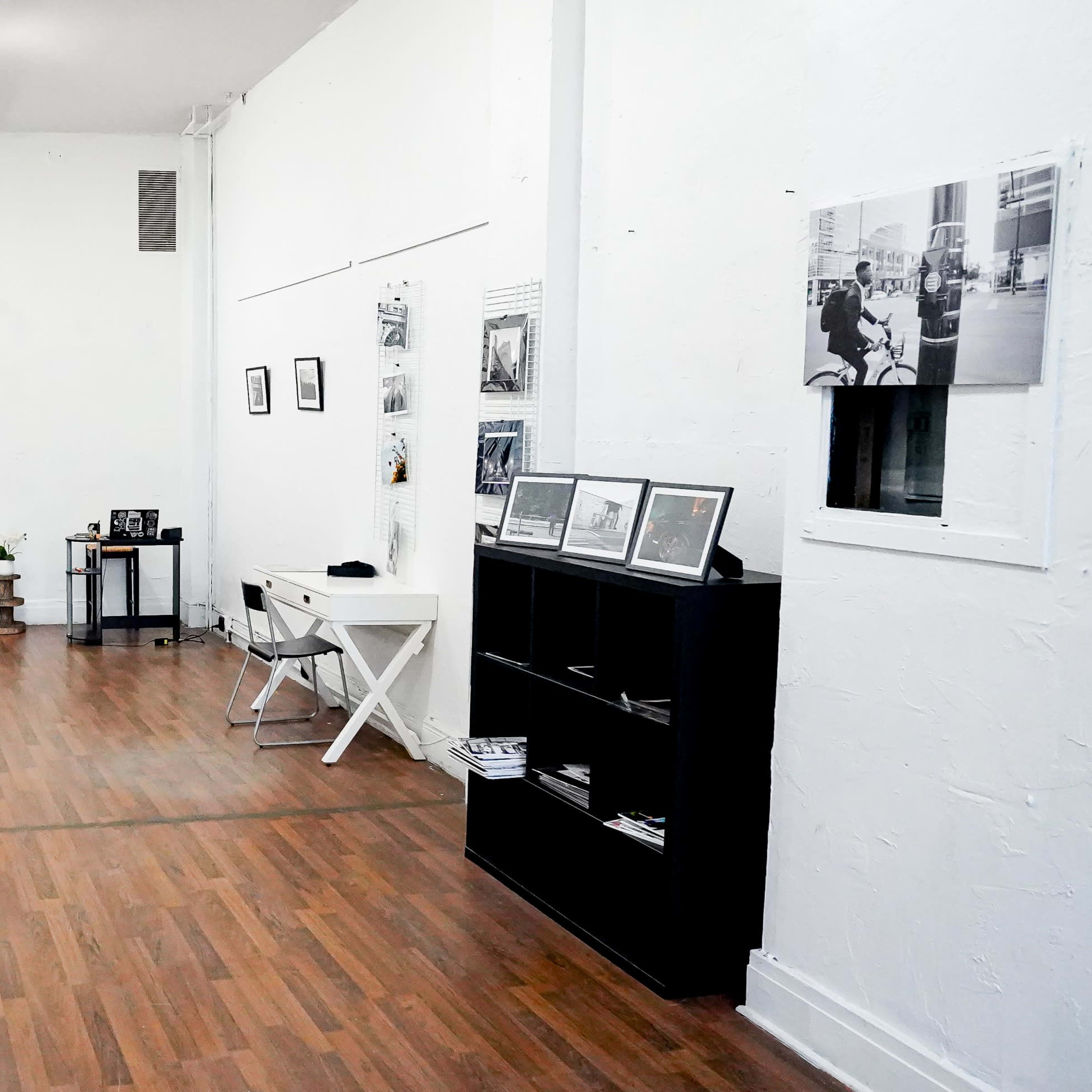 A spacious room with wooden flooring, white walls, and several framed photographs displayed on the walls, along with furniture including tables and a chair.
