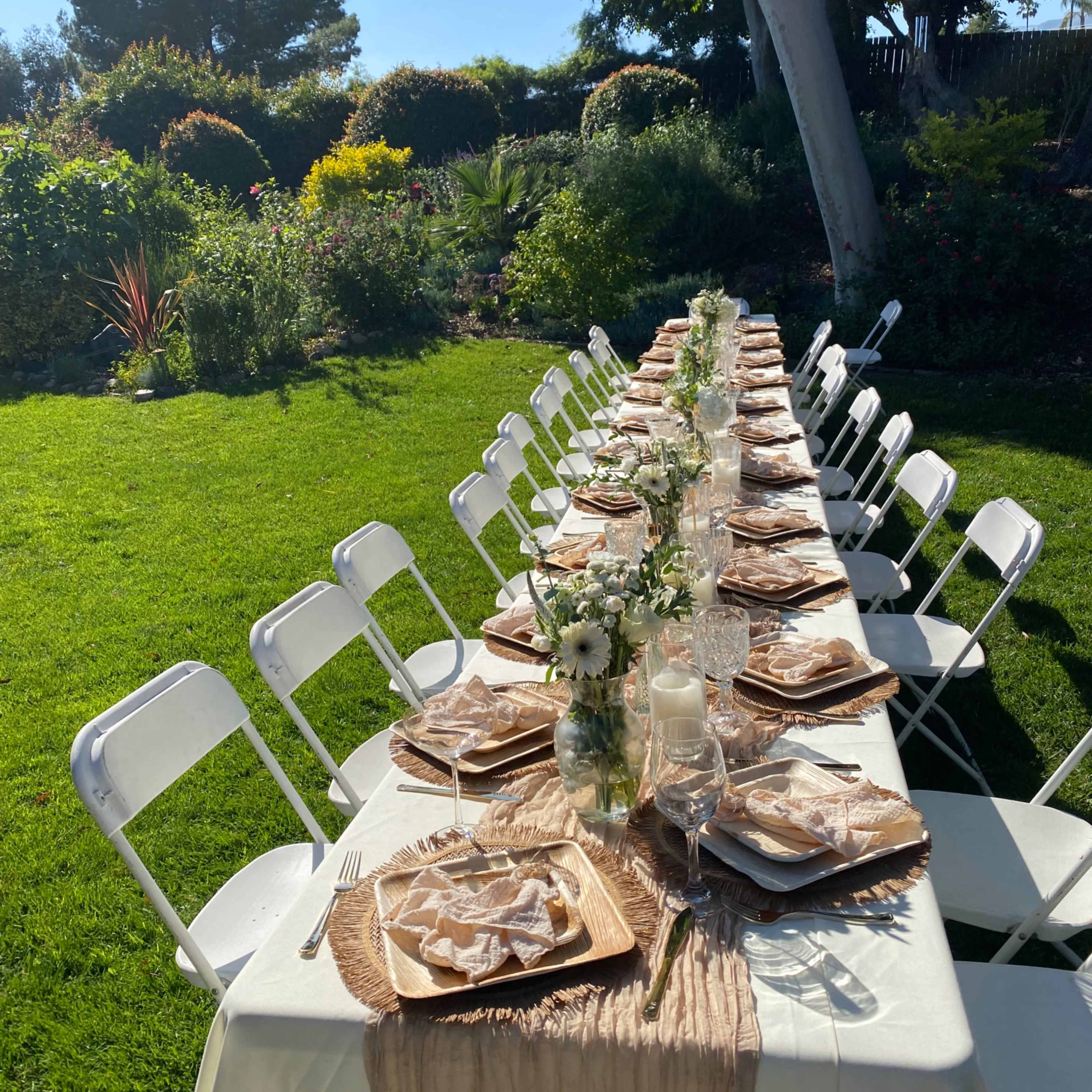 A long, elegantly set table with white chairs is arranged outdoors on a green lawn, surrounded by lush plants and trees.