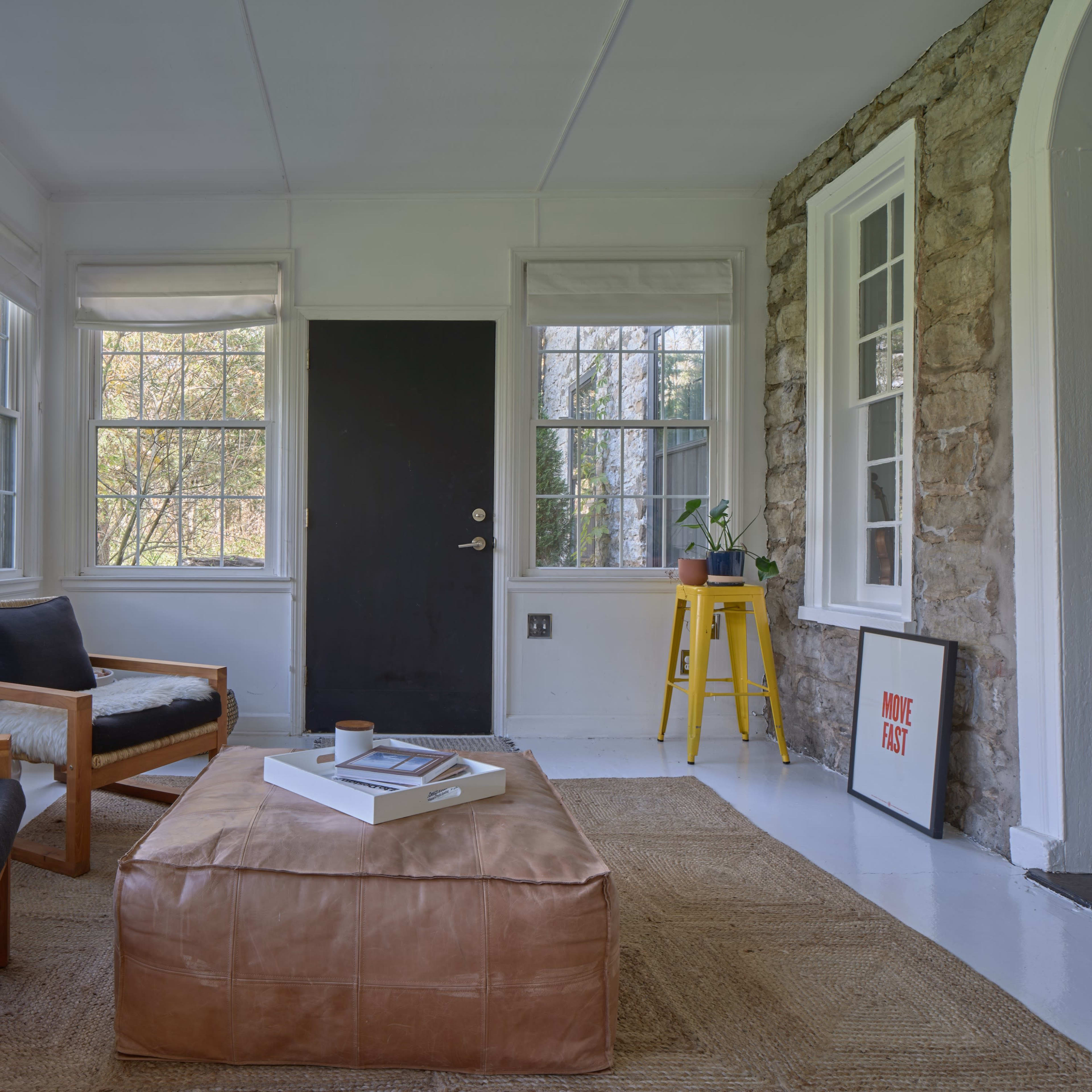 A sunlit room with large windows, featuring two wooden armchairs, a coffee table on a woven rug, and a door leading outside.