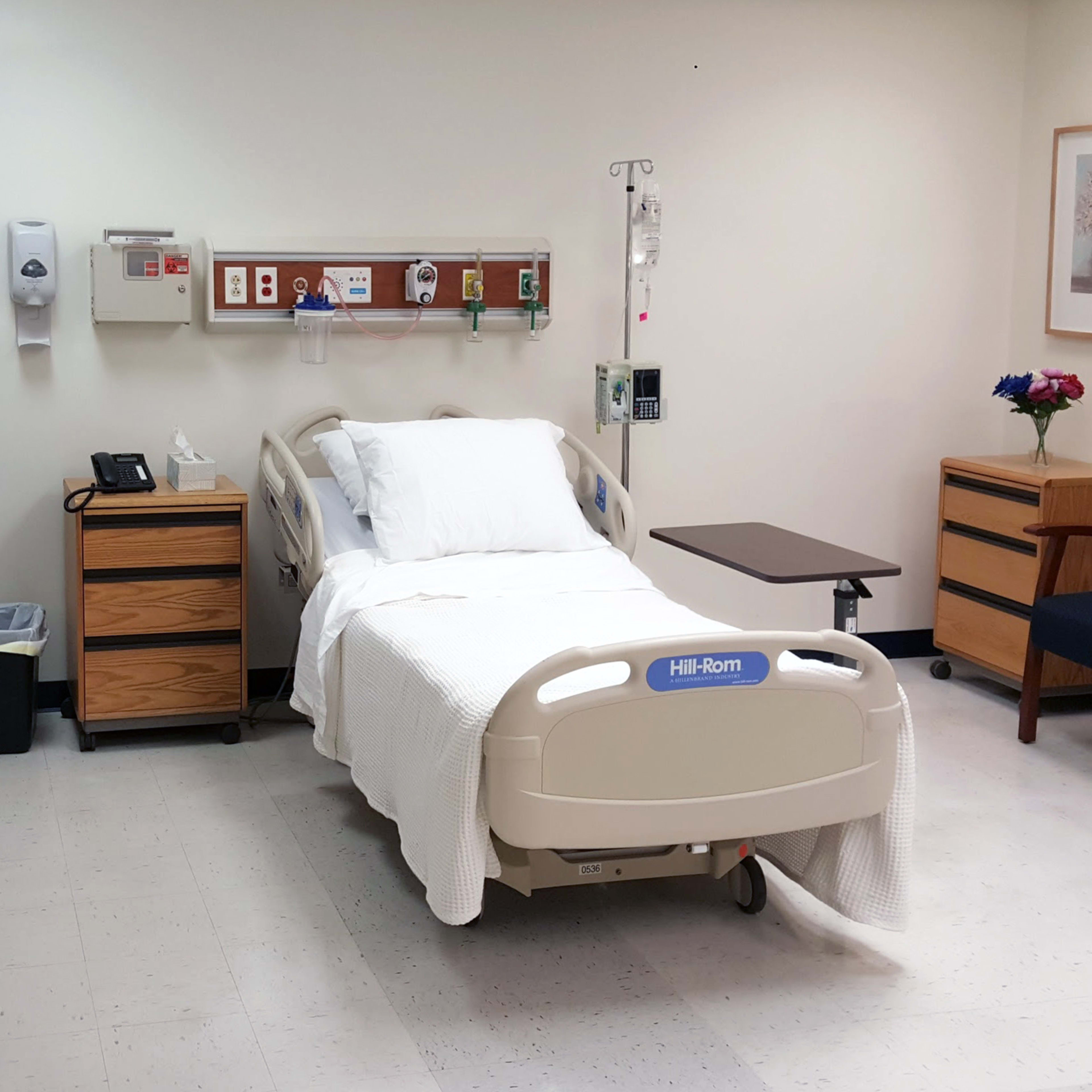 A hospital room featuring a bed with white linens, a bedside table, a chair, and a small bouquet of flowers on a dresser.