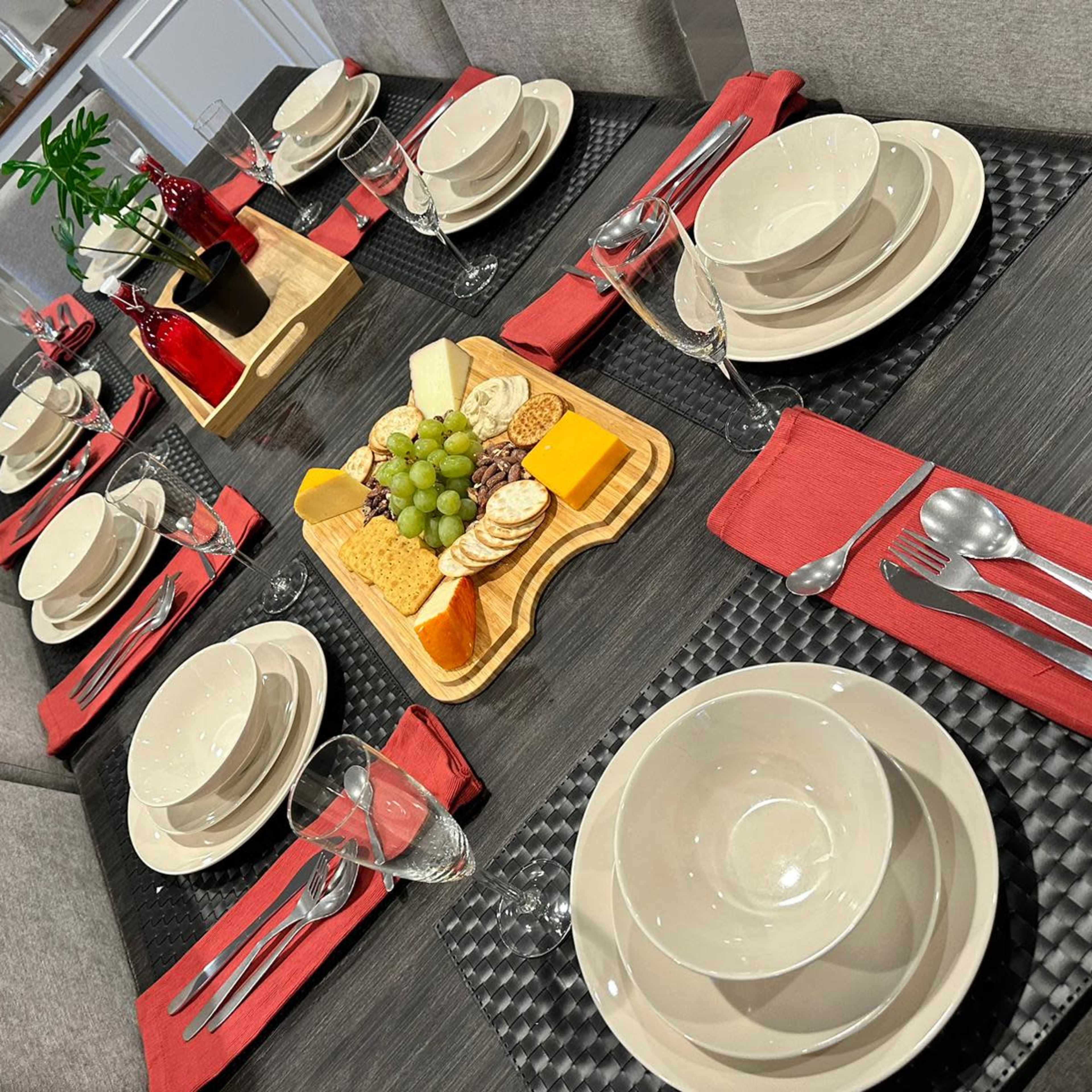 A dining table is set with plates, utensils, glasses, and a wooden platter of cheese and crackers, surrounded by red napkins.
