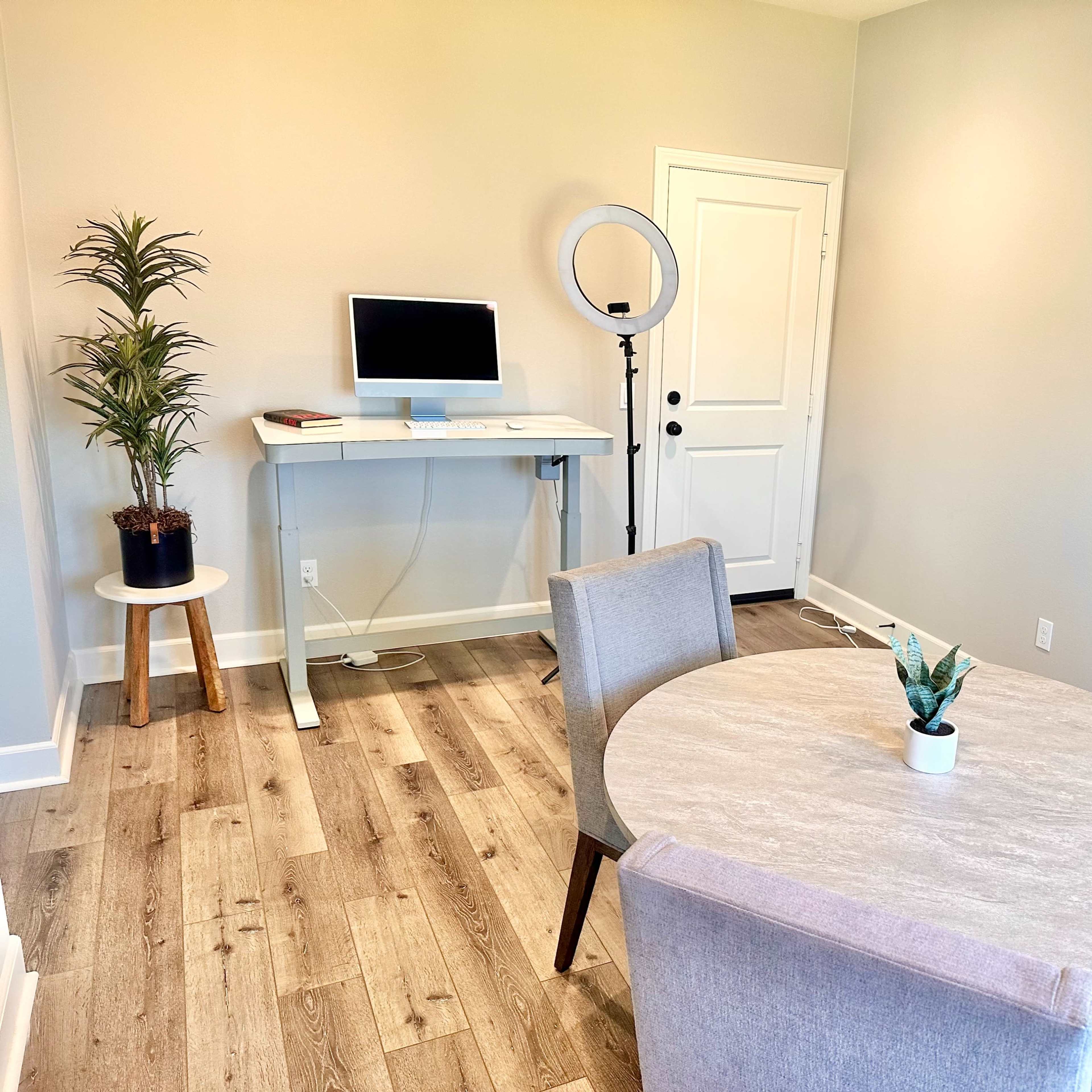 A modern, minimalistic room features a round dining table with two chairs, a small plant centerpiece, a desk with a computer, and a ring light setup beside a door.