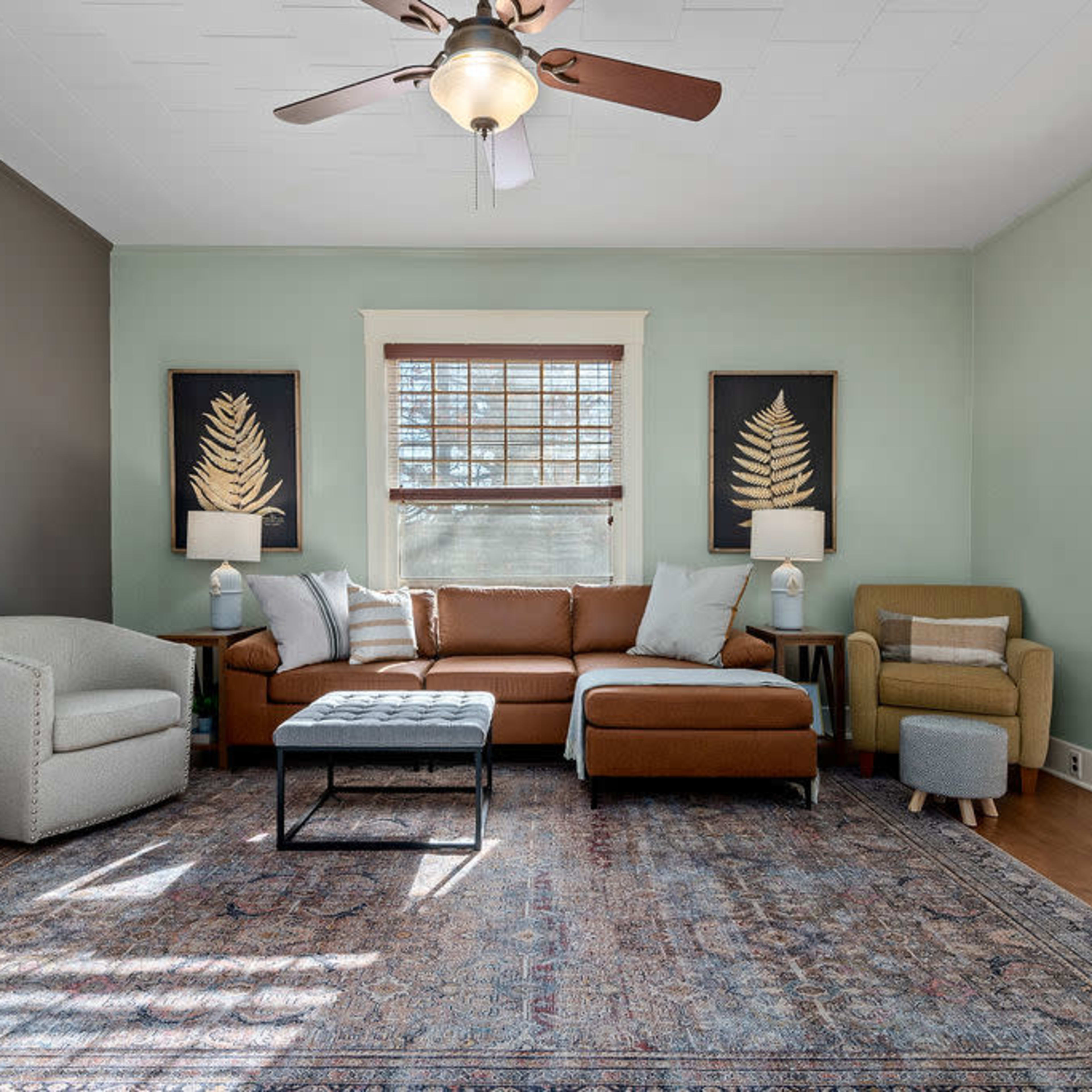 A living room featuring a brown sectional sofa, a gray armchair, and a patterned area rug, with wall art and large windows providing natural light.