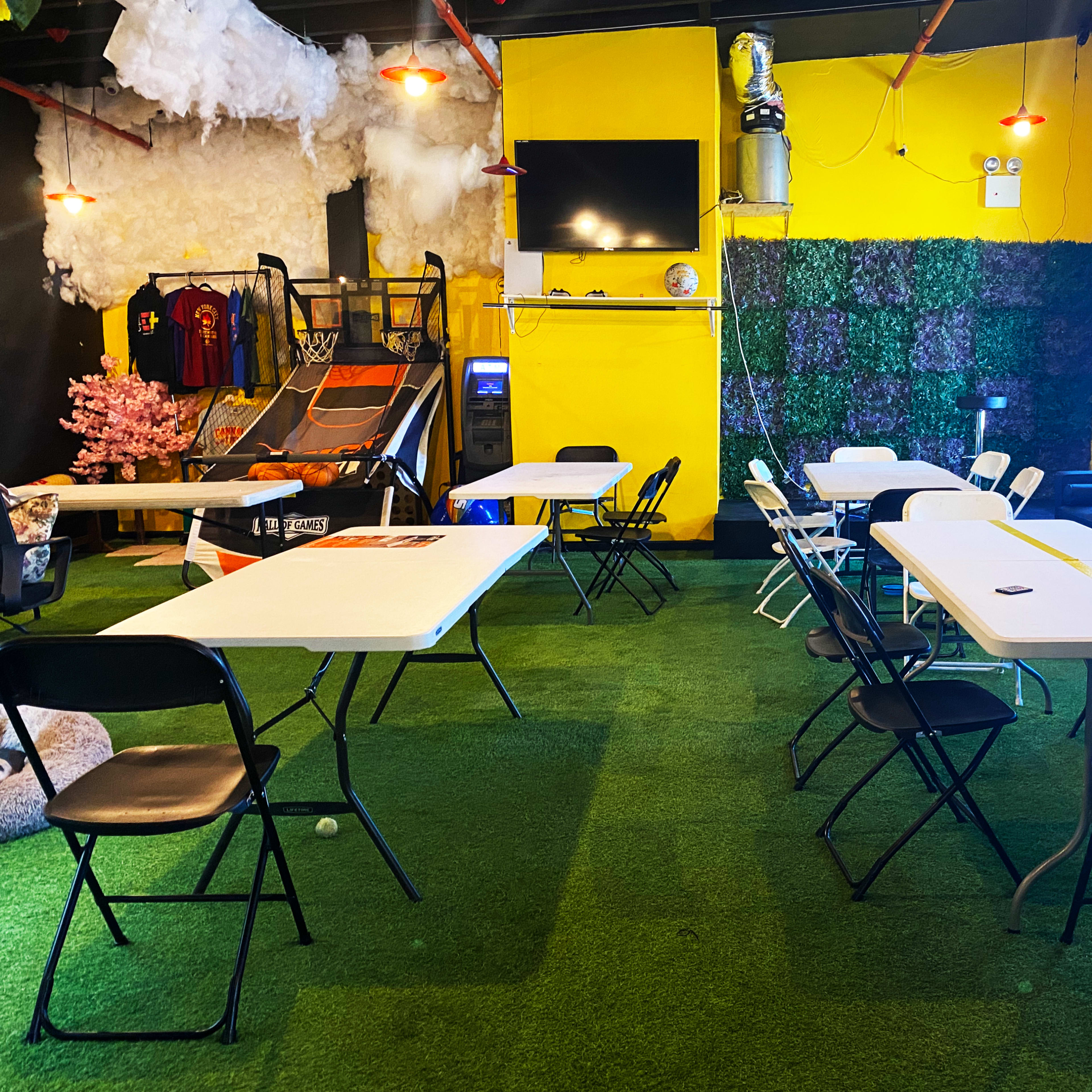 The scene features a brightly colored room with artificial grass flooring, several tables and chairs arranged around, a basketball hoop, and a large screen mounted on the wall.