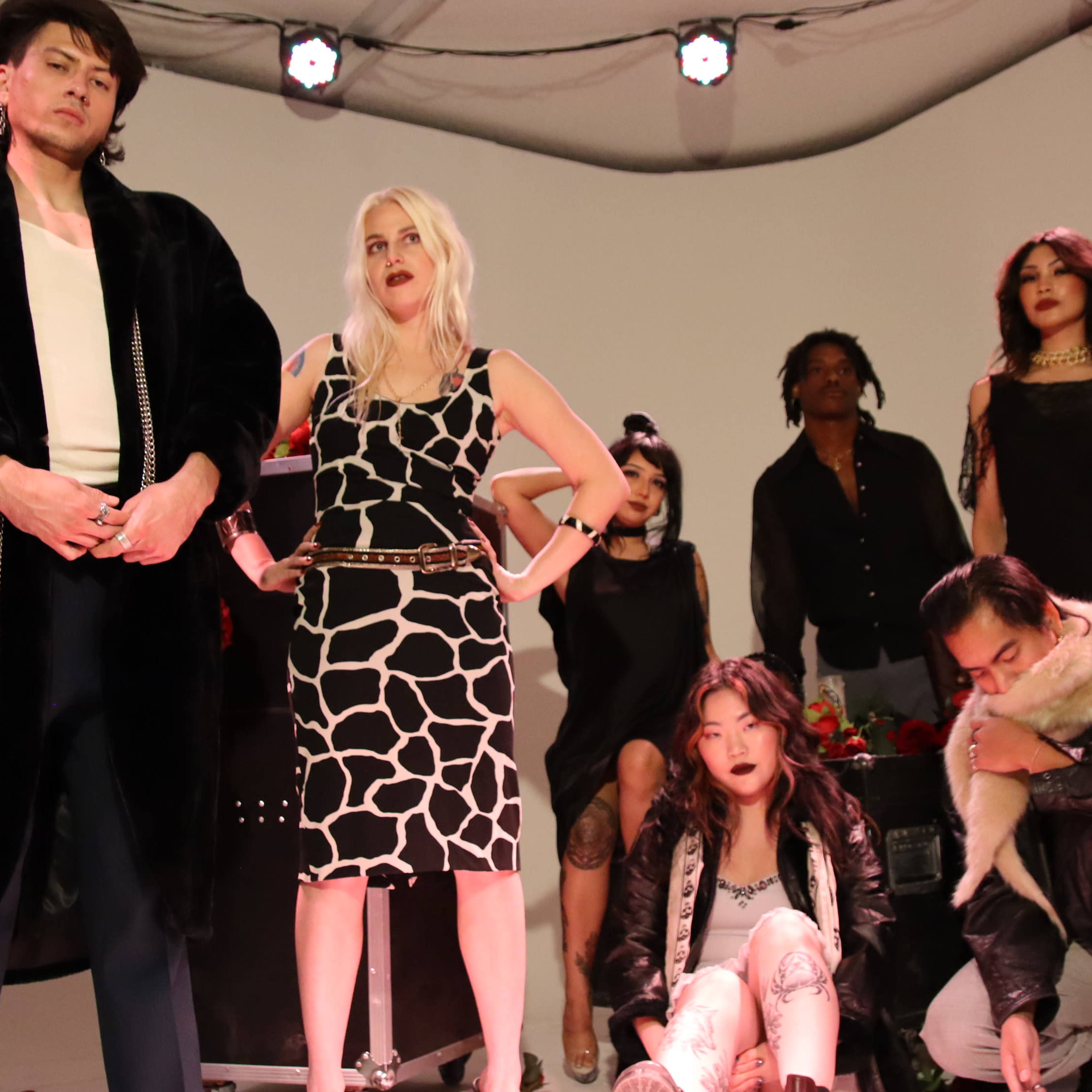 A group of fashion models poses on a stage with various outfits, featuring a mix of standing and seated positions against a simple backdrop.