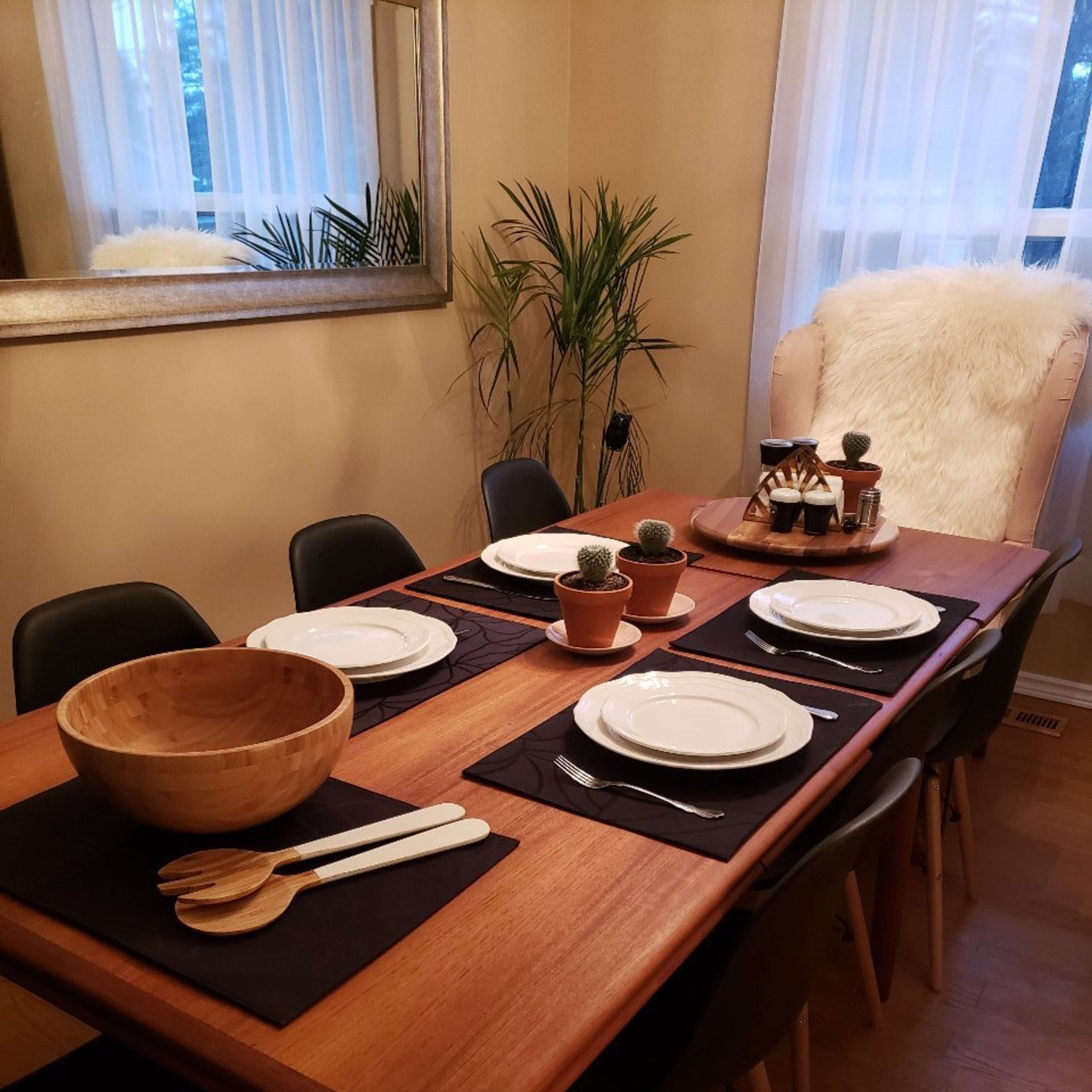 A wooden dining table is set with white plates and utensils, surrounded by black chairs, with a bowl and potted plants as centerpieces.