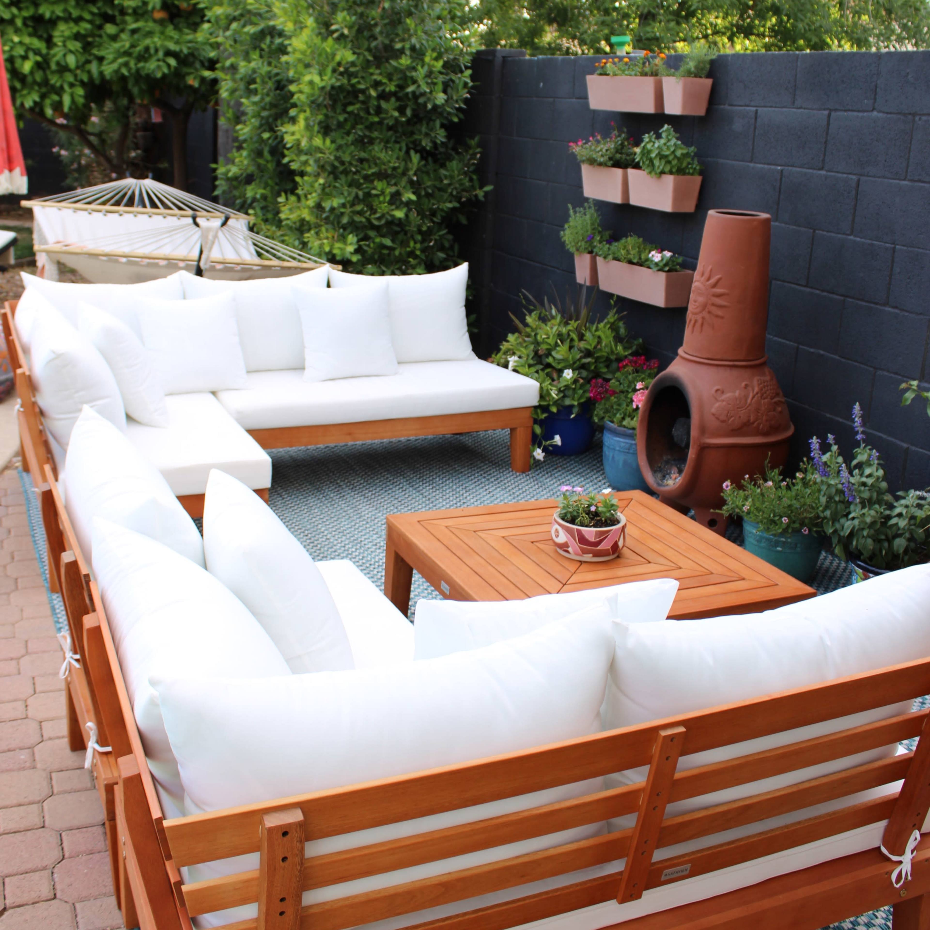 A cozy outdoor seating area arranged with white cushions on wooden furniture, a coffee table at the center, and planters with greenery along the walls.