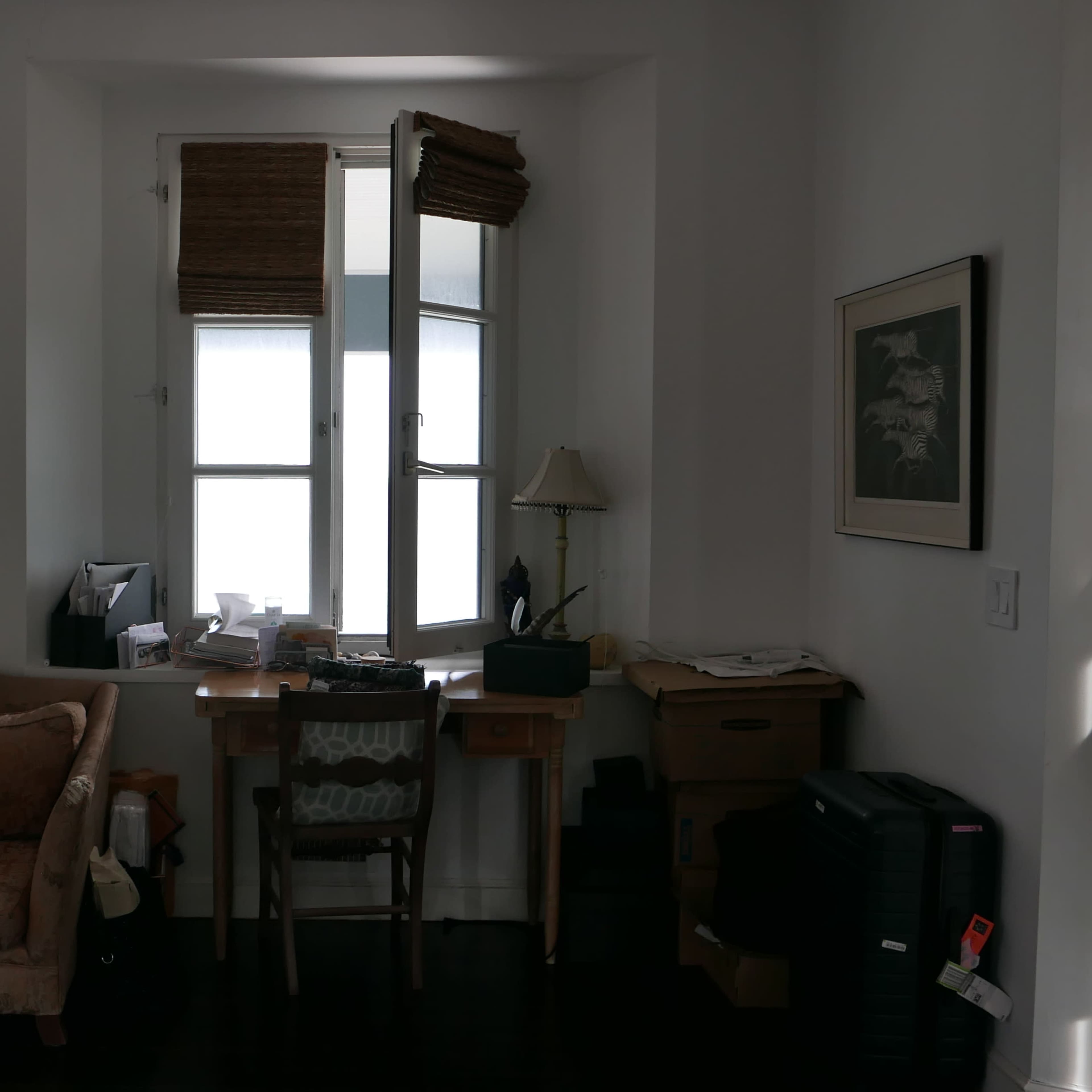 A sofa is placed near a window, alongside a small desk and various items in a well-lit room with white walls.