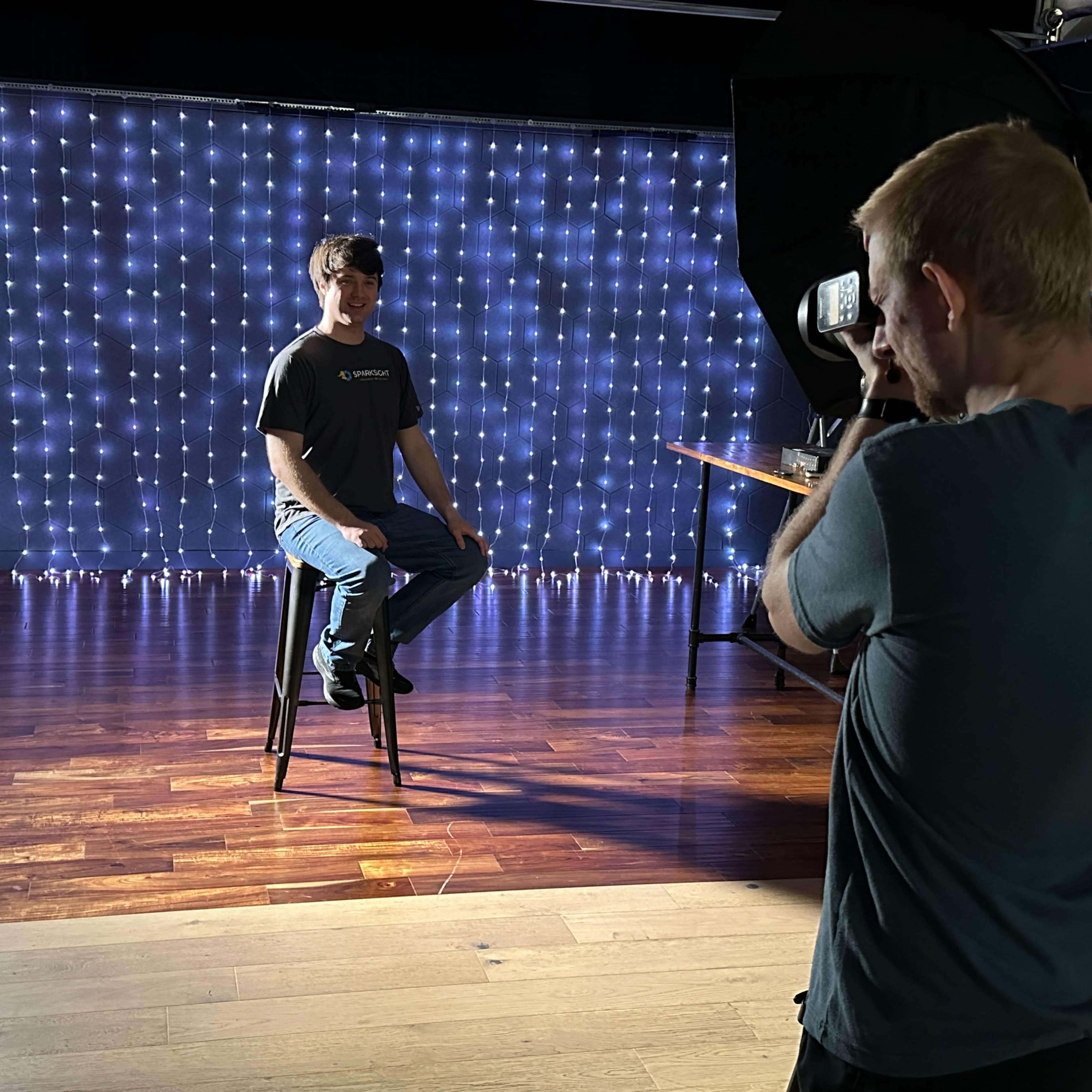 A person sits on a stool in front of a backdrop of string lights while another person films with a camera.
