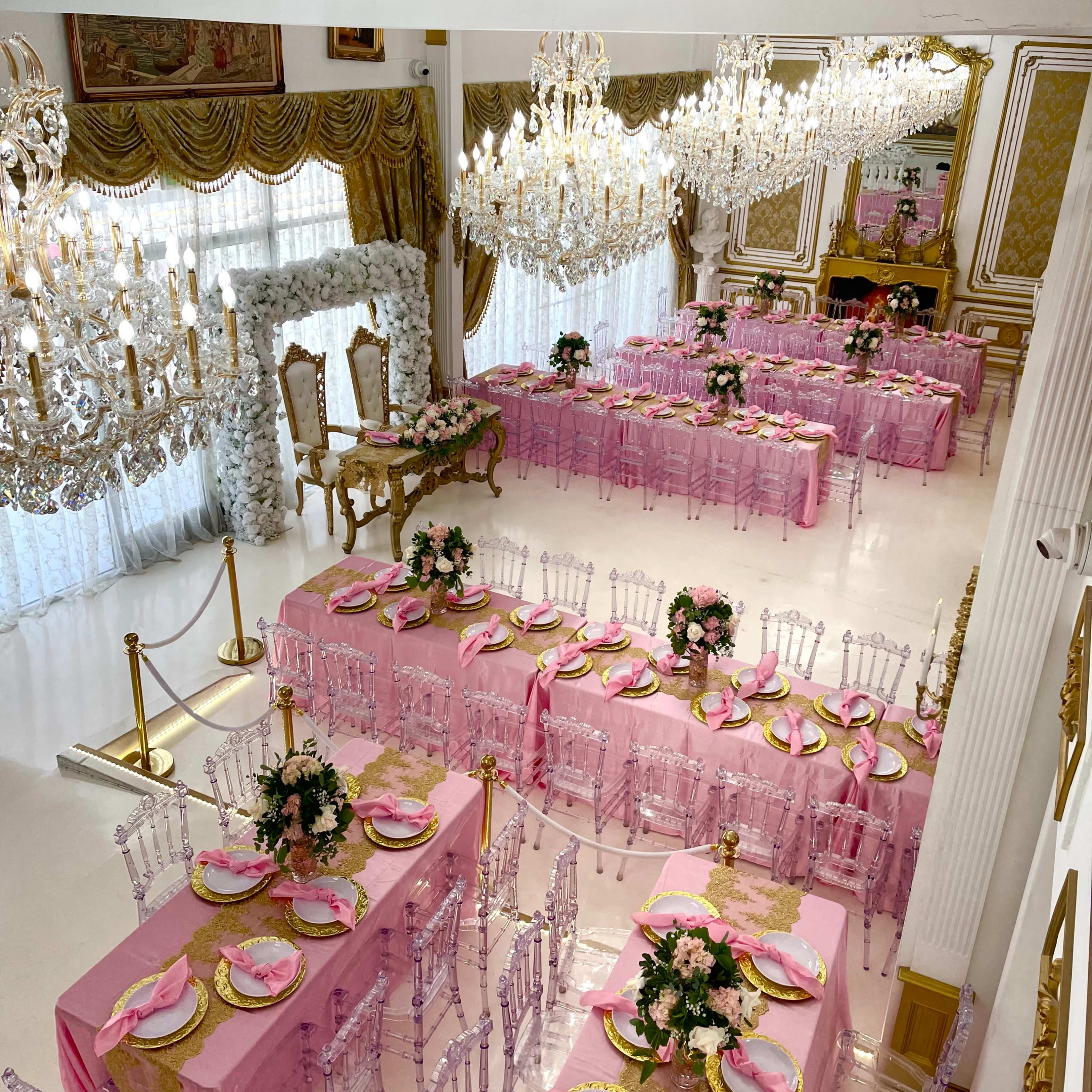 A lavish banquet hall set up for an event, featuring pink tablecloths, clear acrylic chairs, and ornate chandeliers.