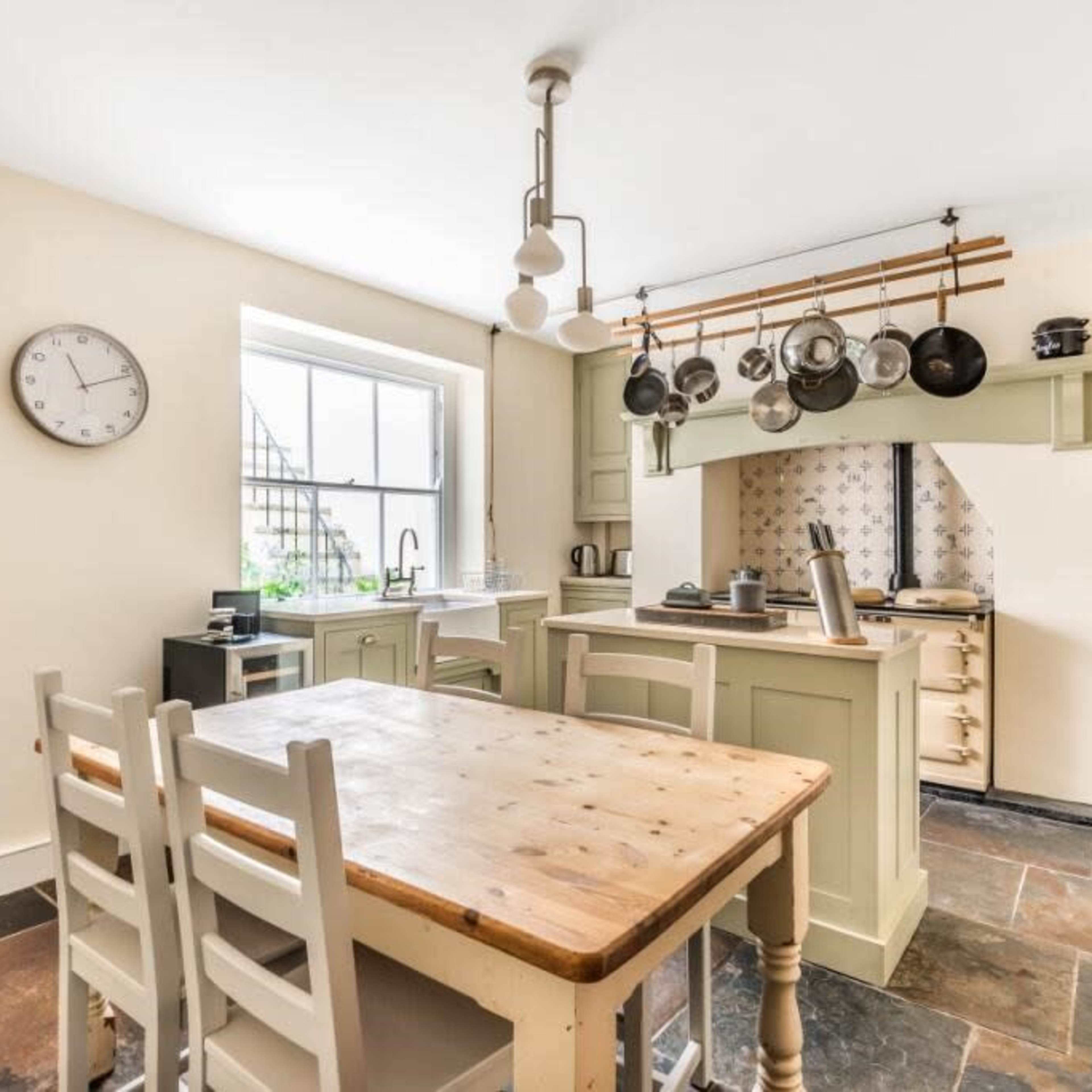 A kitchen with light-colored walls, a wooden dining table, and pots hanging from a rack above the stove.
