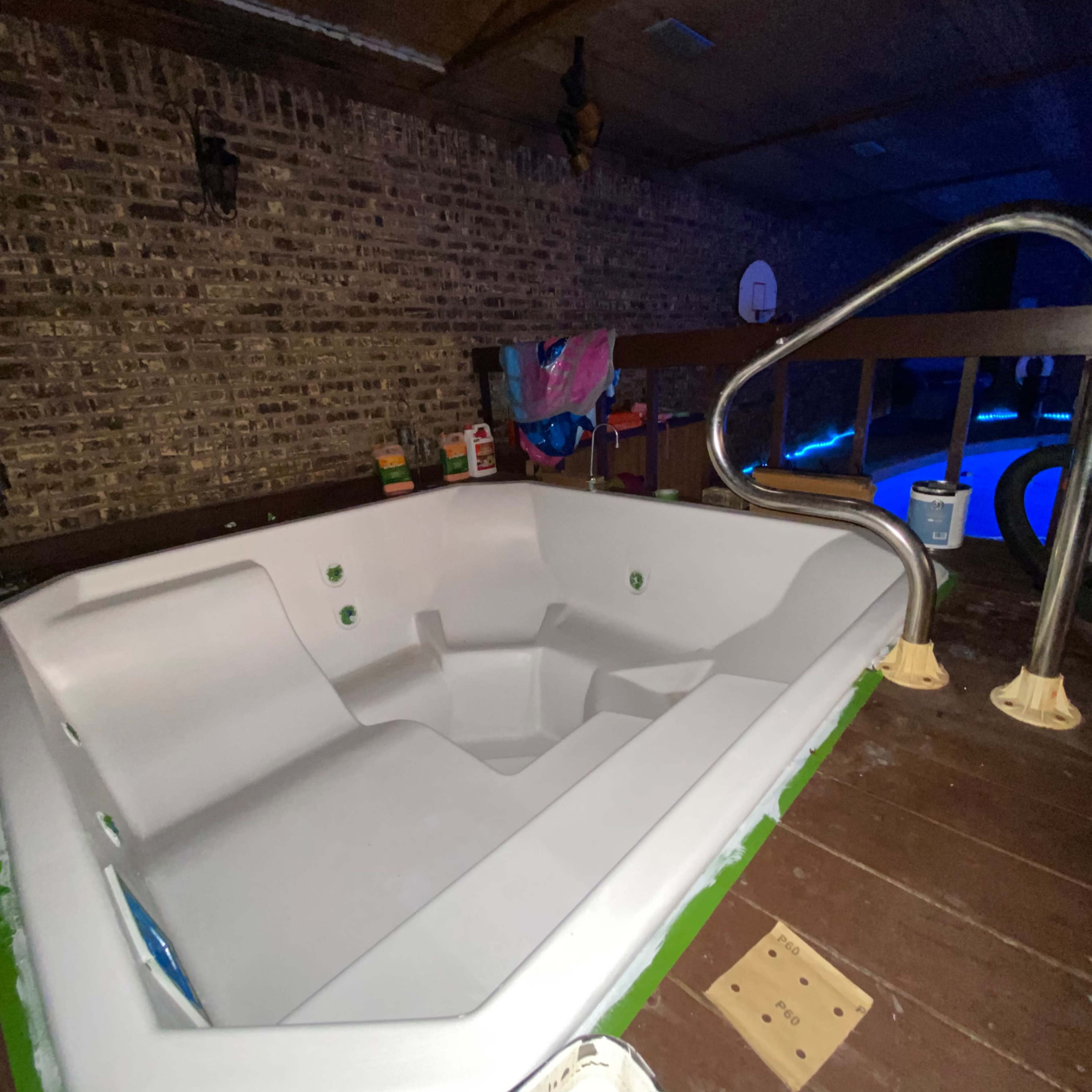 A fiberglass hot tub is situated in a dimly lit indoor space with wooden flooring and a brick wall backdrop.