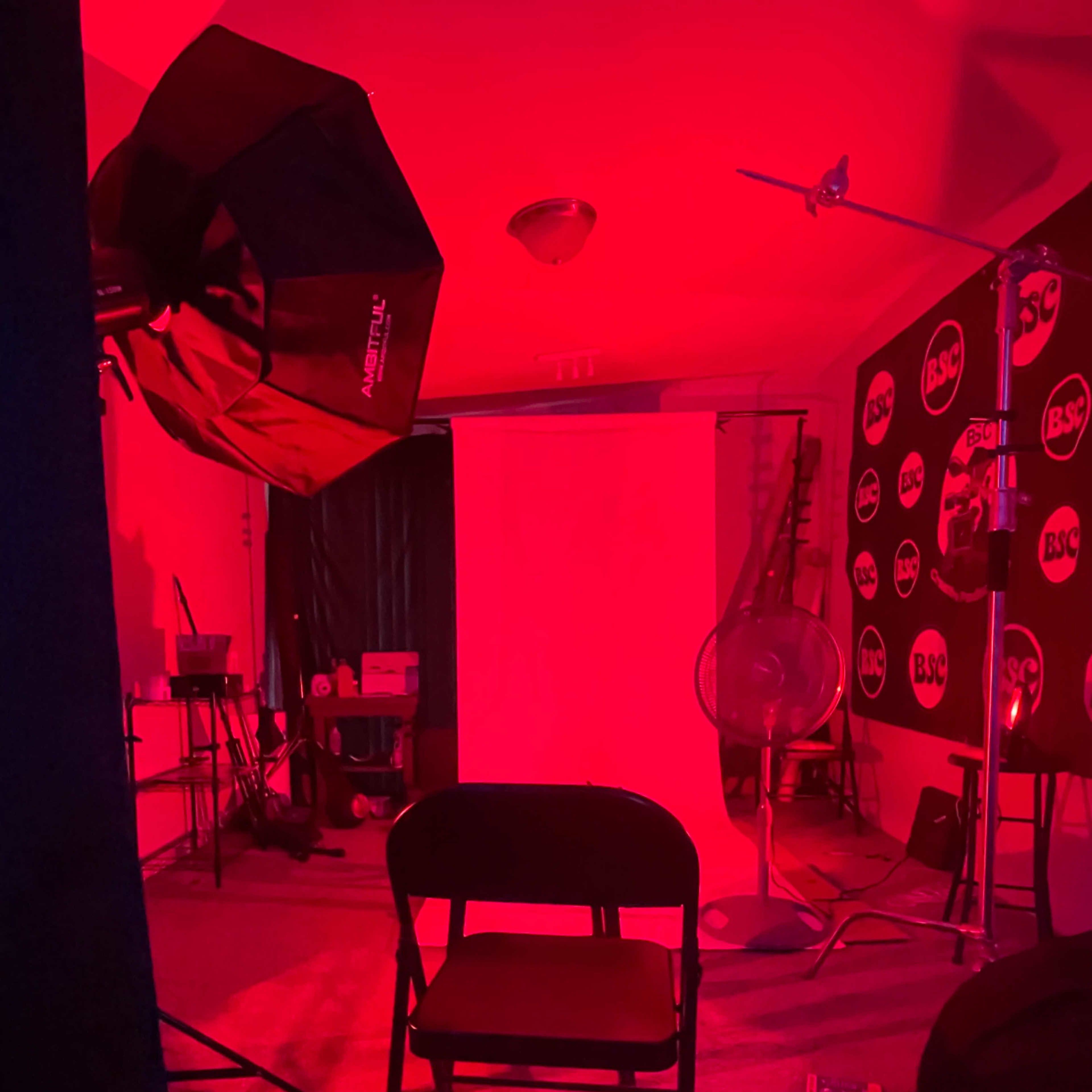 A room with a red-lit backdrop, a black folding chair, and studio lighting equipment.