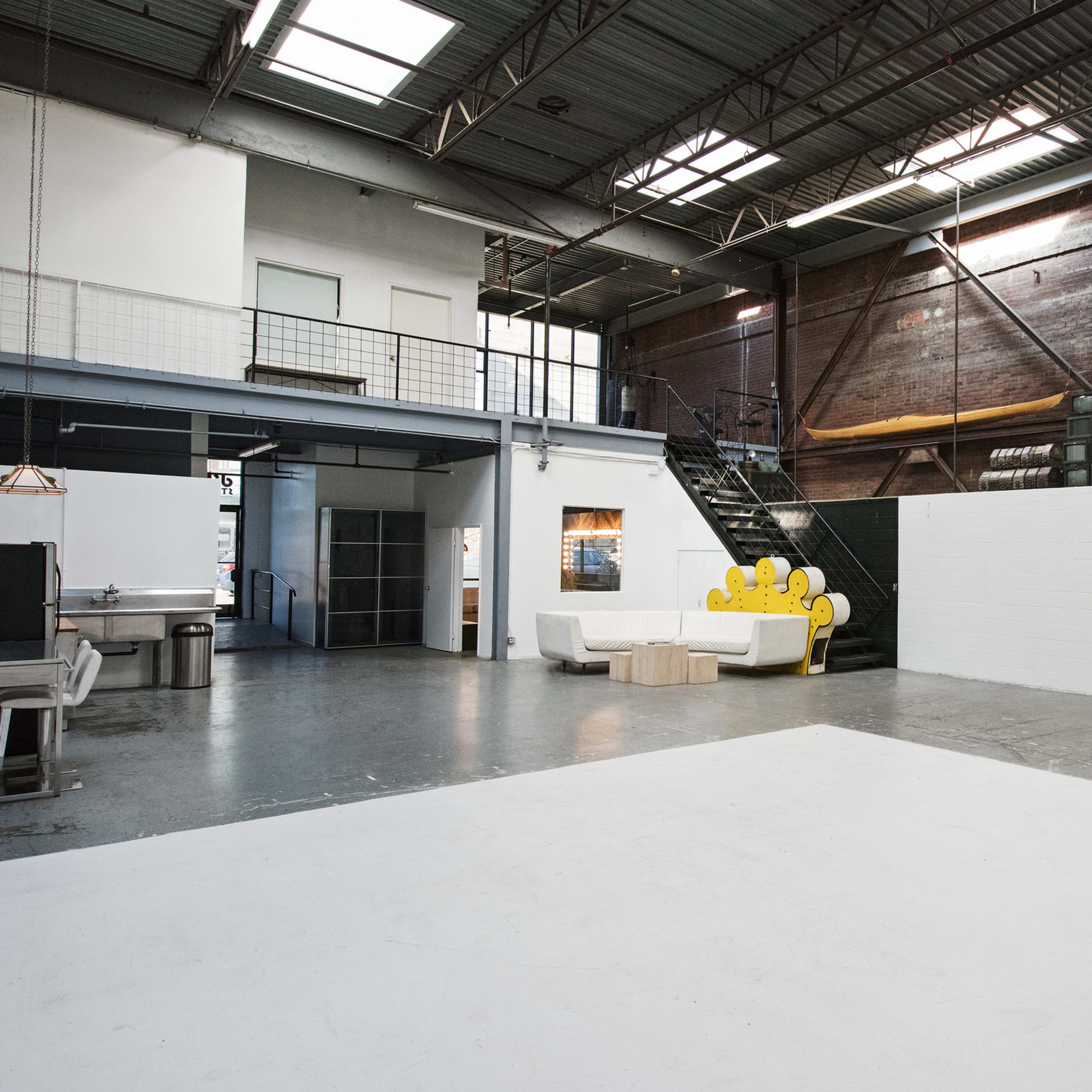 A spacious industrial interior with an open layout, featuring a white wall, a staircase, and a yellow seating area.