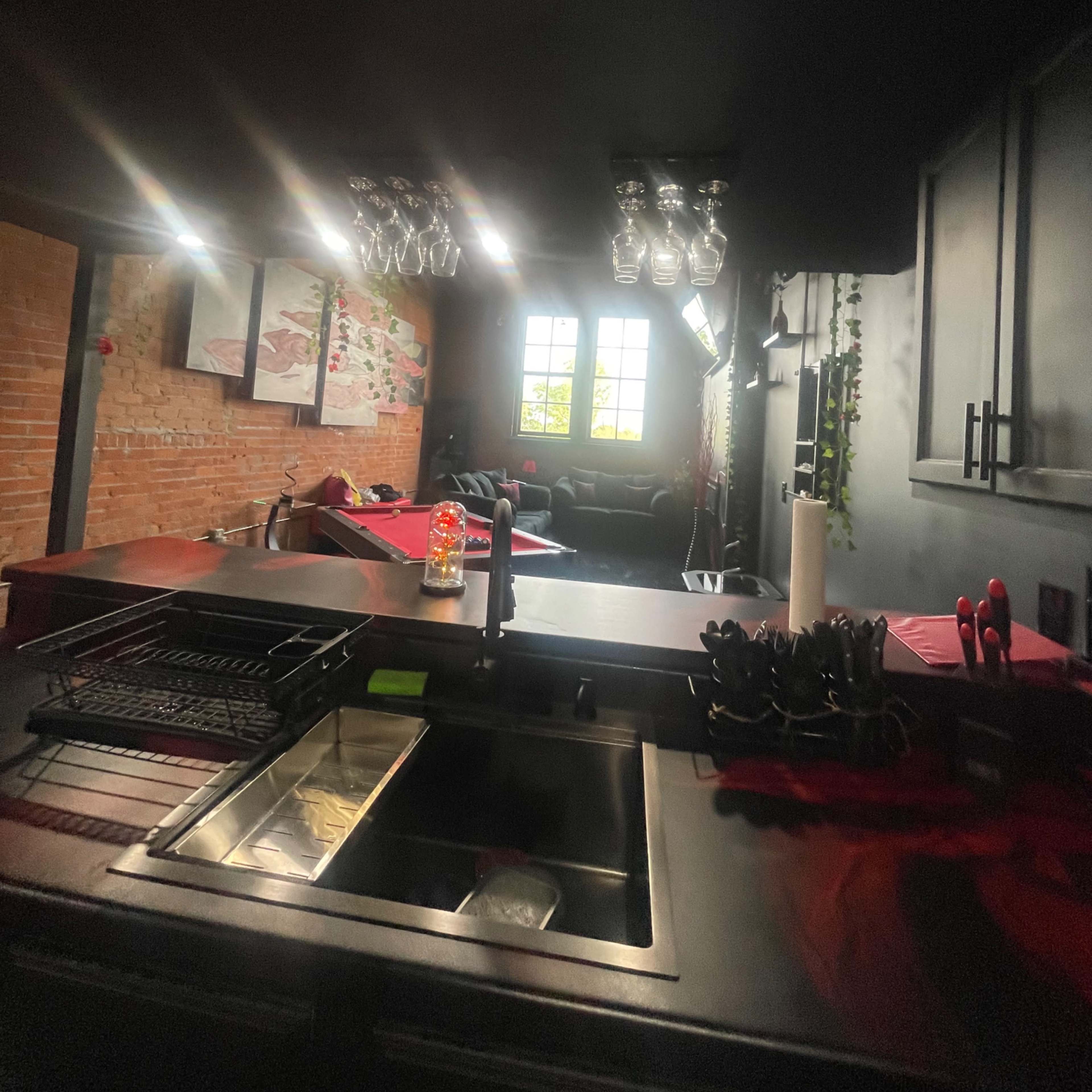 A modern kitchen with a black ceiling, hanging glassware, and a window revealing a living area.