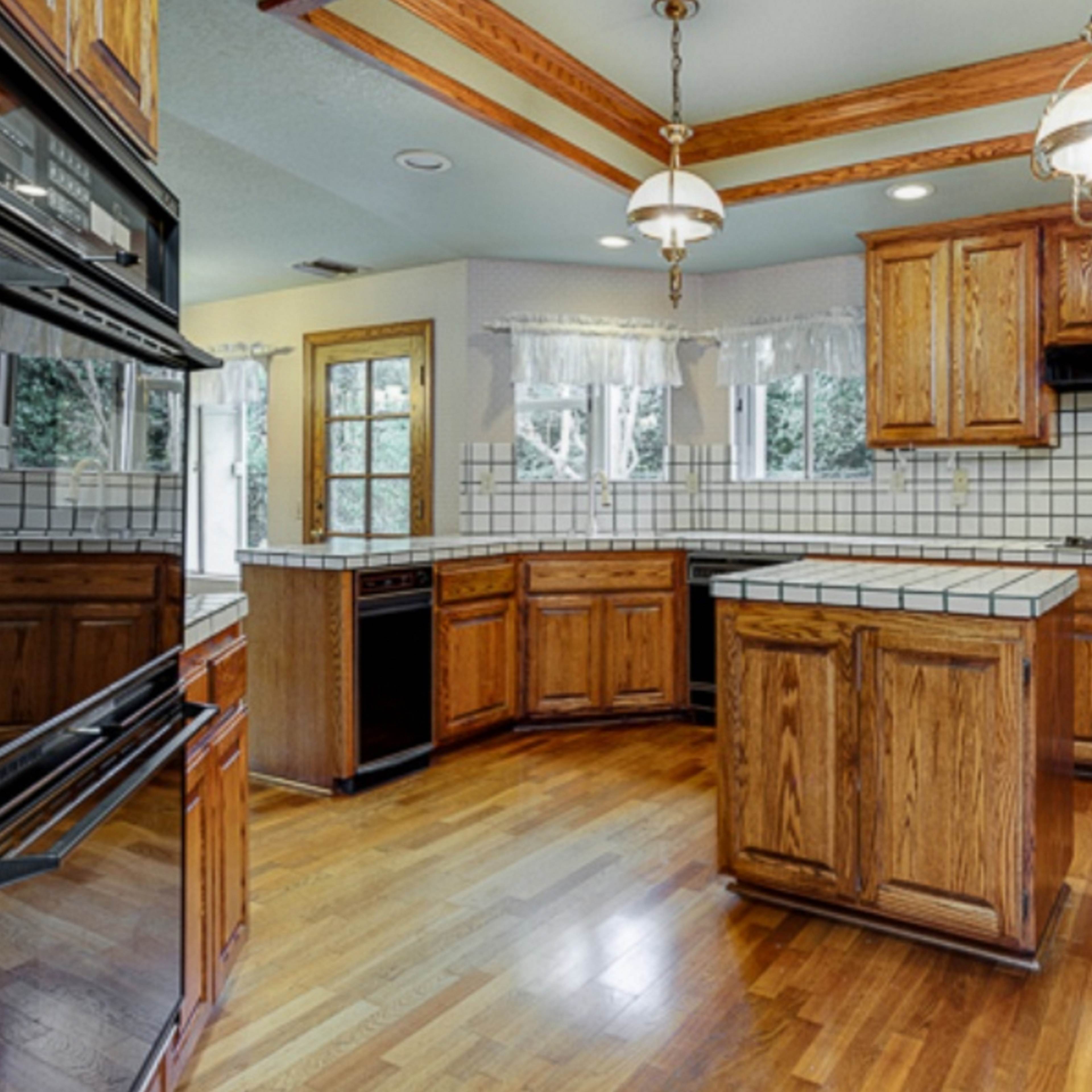 A kitchen features wooden cabinets, a tiled island, and appliances against a backdrop of large windows.