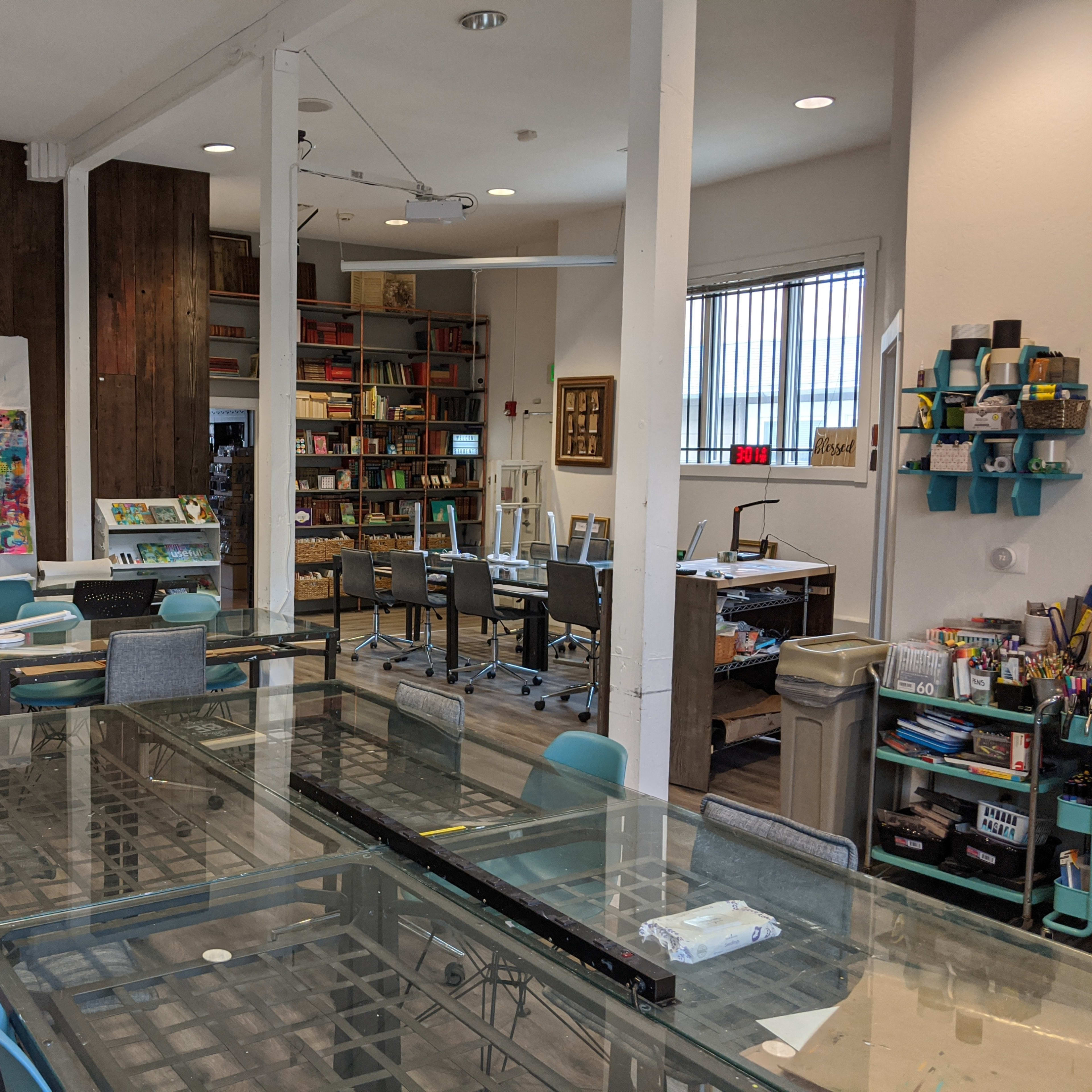 A bright art studio with tables, chairs, and shelves filled with various art supplies and books.