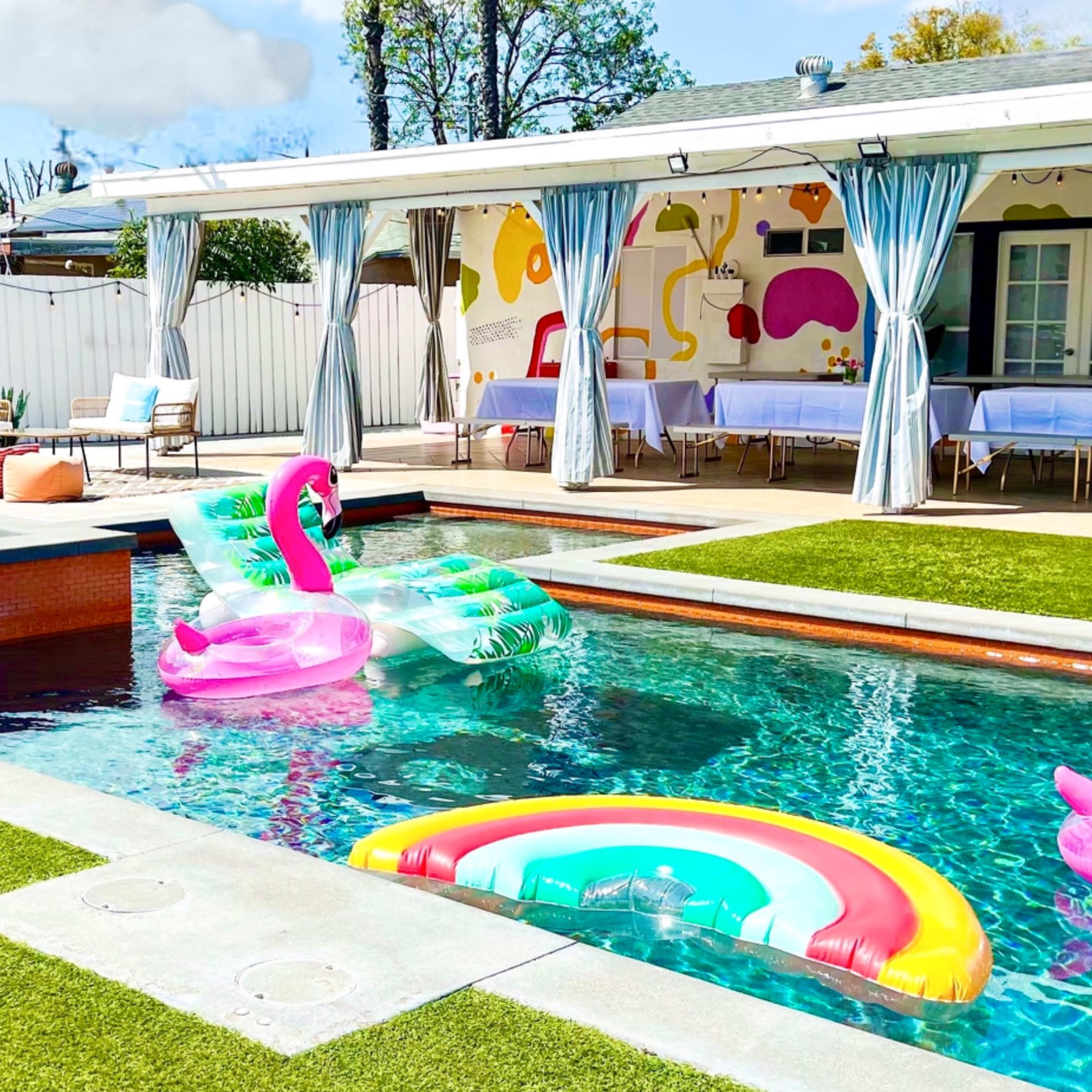 A vibrant backyard pool area featuring colorful inflatable floats, a covered seating area, and freshly manicured grass.